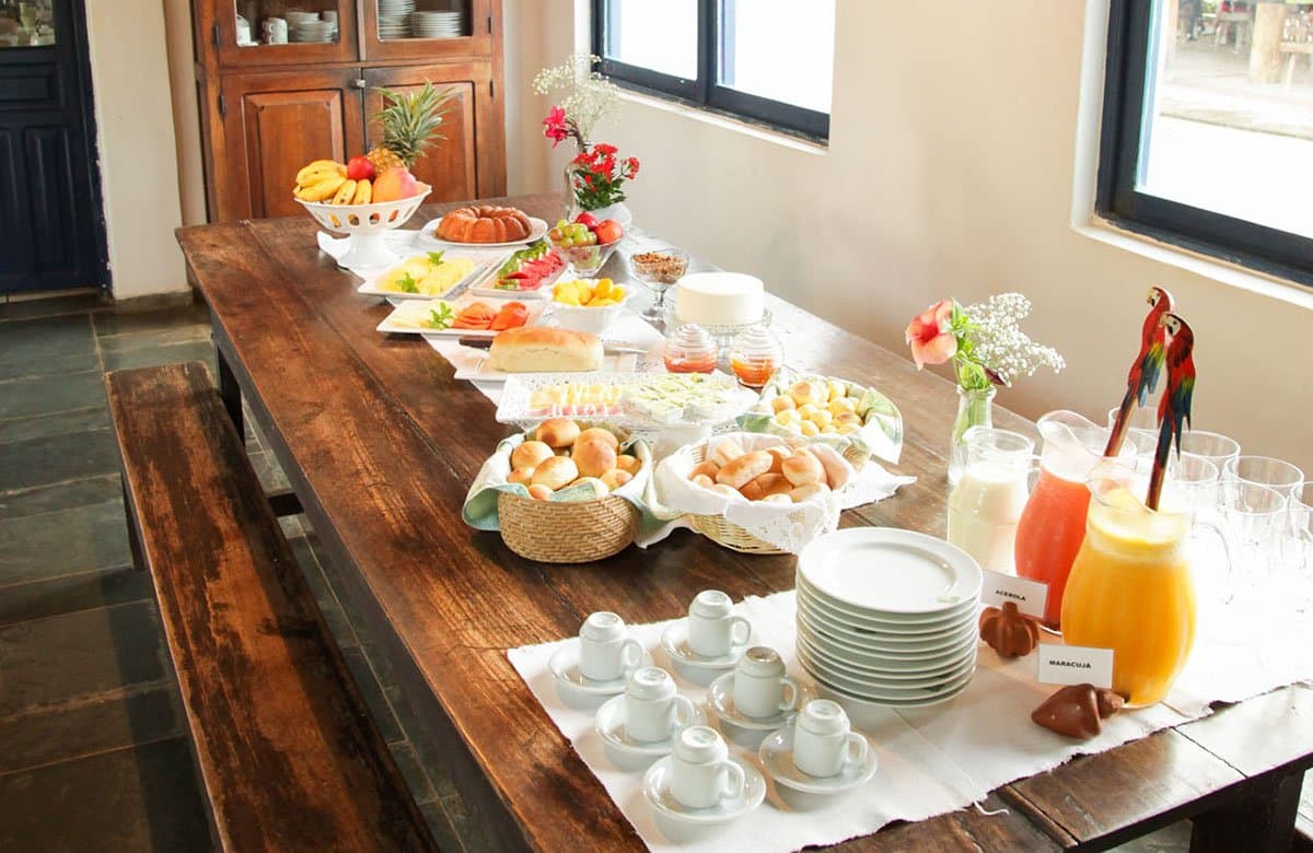 Fresh fruit, warm bread rolls, and tropical juices — fuel for a full day of wildlife adventure. Photo: Alexis Prappas - www.prappas.com A generous Brazilian breakfast buffet laid out on a long wooden table at a Pantanal tour from Campo Grande lodge, featuring fresh tropical fruits, bread rolls, cakes, acerola and maracujá juices, and small espresso cups