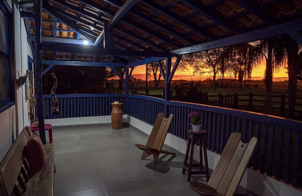 Goed gezelschap begint op een veranda als deze. Overdekt terras met blauw geschilderde houten reling en rustieke stoelen met uitzicht op een wijd open landschap in een dieporanje zonsondergang tijdens een Pantanal-tour vanuit Campo Grande