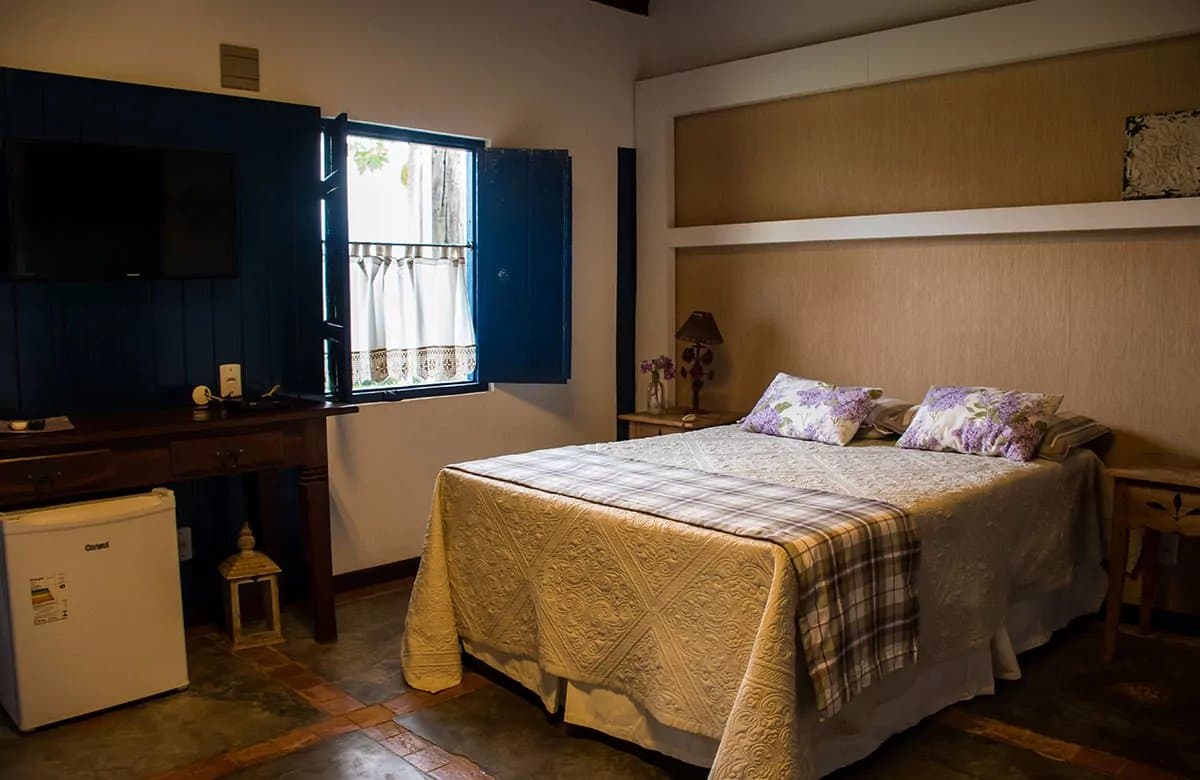 Rustig slapen na een dag vol wilde dieren. Gezellige tweepersoonskamer van een lodge voor een Pantanal-tour vanuit Campo Grande, met gewatteerde sprei, paarse bloemensierkussens, blauwe houten luiken open naar tropisch groen en een houten bureau