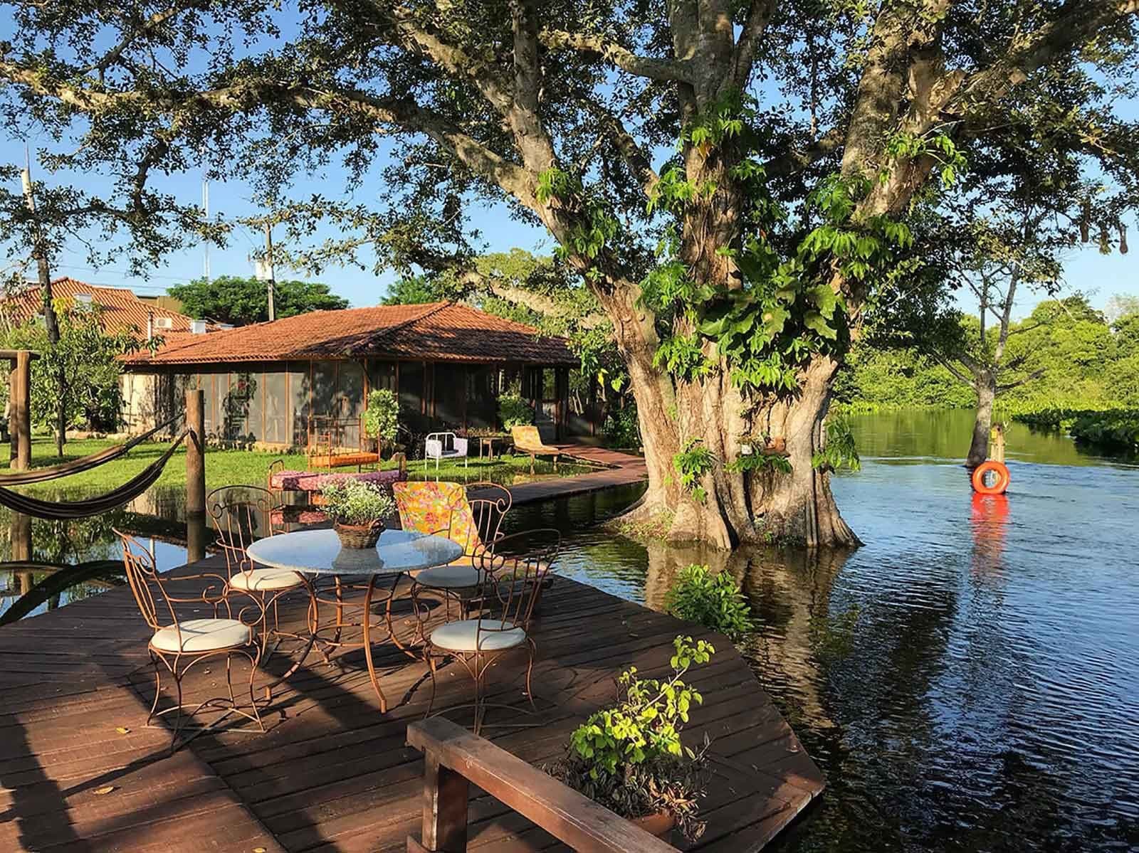 Dit is het soort plek waar je agenda vanzelf verdwijnt. Een houten terras met smeedijzeren stoeltjes kijkt uit over kalm rivierwater; een grote boom groeit aan de oever en een ecolodge met terracotta dak is zichtbaar achter weelderig groen