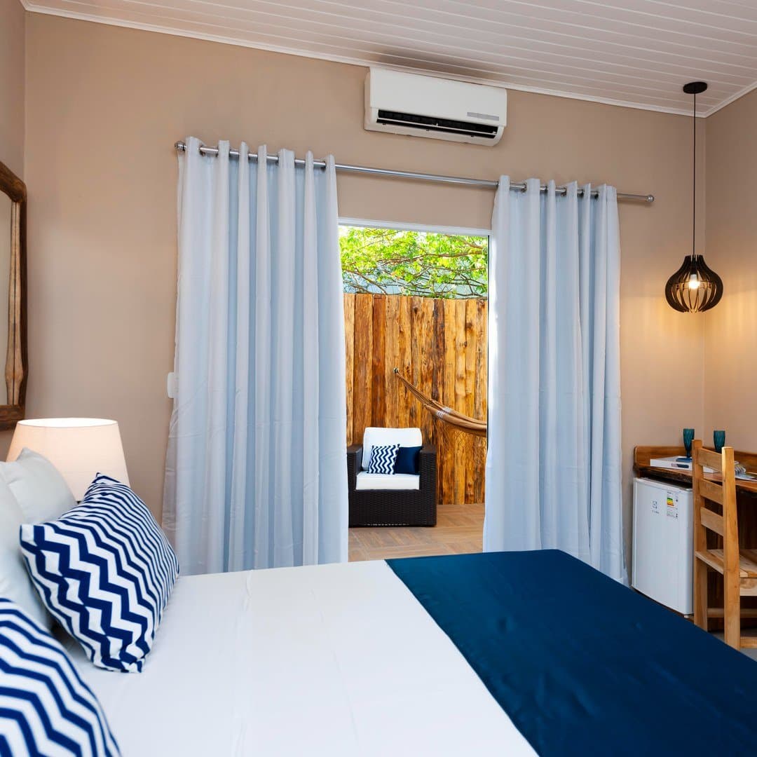 Komfort und Hängematte — die perfekte Erholung nach einem Abenteuertag. Klimatisiertes Doppelzimmer auf einer Pantanal-Tour ab Cuiabá, mit marine Kissen im Zickzackmuster, weißen Vorhängen und einer privaten Holzterrasse mit Hängematte und Liegestuhl durch die geöffnete Schiebetür sichtbar