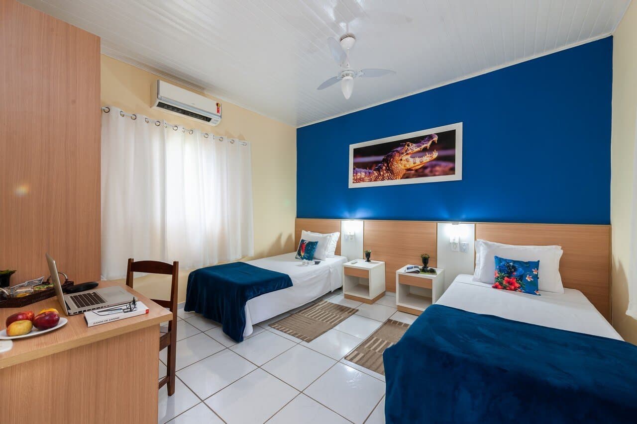 Marineblaue Decken, ein Kaiman an der Wand — Ihr Rückzugsort zwischen zwei Abenteuern. Zweibettzimmer in einer Unterkunft der Pantanal-Tour ab Cuiabá mit marineblauer Bettwäsche, blauer Akzentwand, einem gerahmten Kaimanfoto und einem Schreibtisch mit frischem Obst
