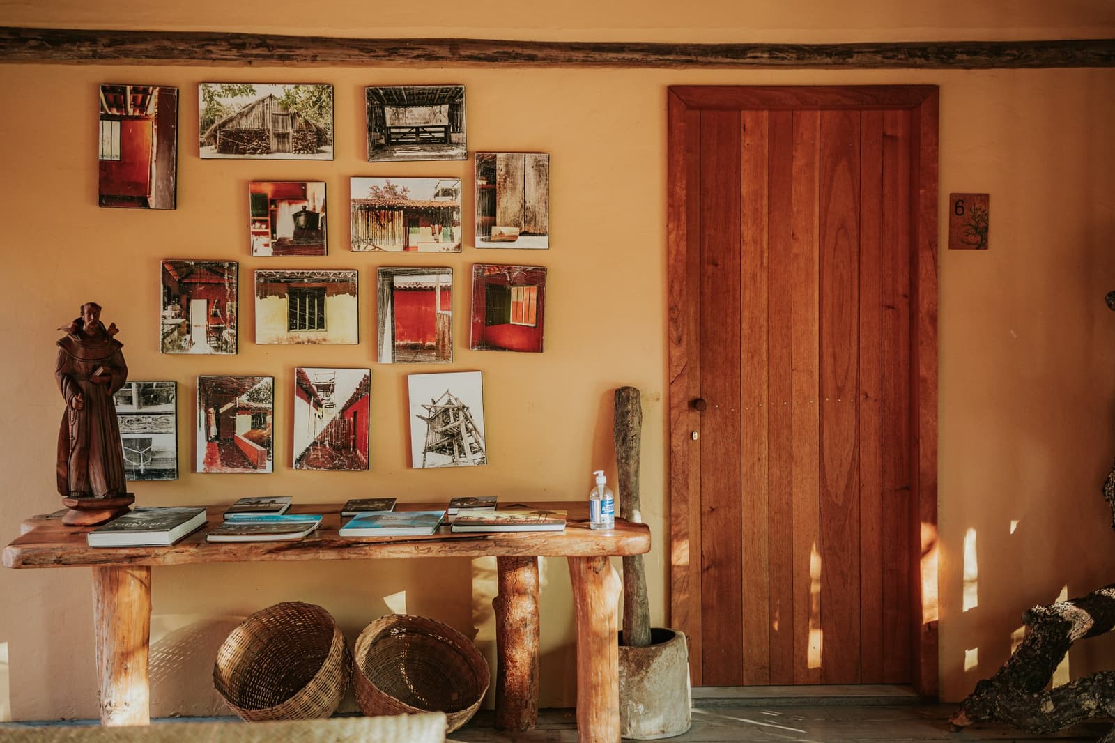 Handcrafted details and local stories lining the walls of Pousada Trijunção. Photo: Albori Ribeiro Warm ochre wall at Pousada Trijunção decorated with a grid of photographic prints depicting traditional Cerrado architecture, beside a handcrafted wooden console table holding books, a carved monk figurine and woven baskets
