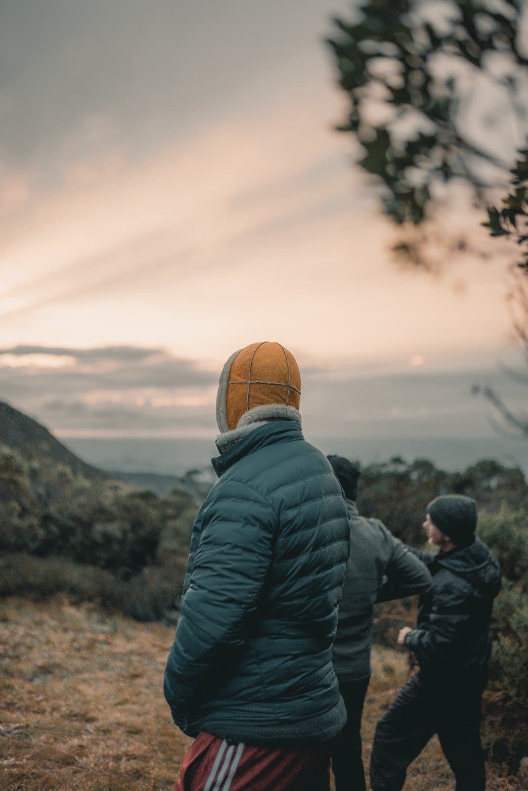 First light over the Mantiqueira — worth every cold, early morning. Two hikers in heavy winter layers and a sheepskin hat stand on an exposed ridge during Serra Fina Trekking, gazing over misty mountain valleys beneath a soft pastel sunrise sky in the Serra da Mantiqueira