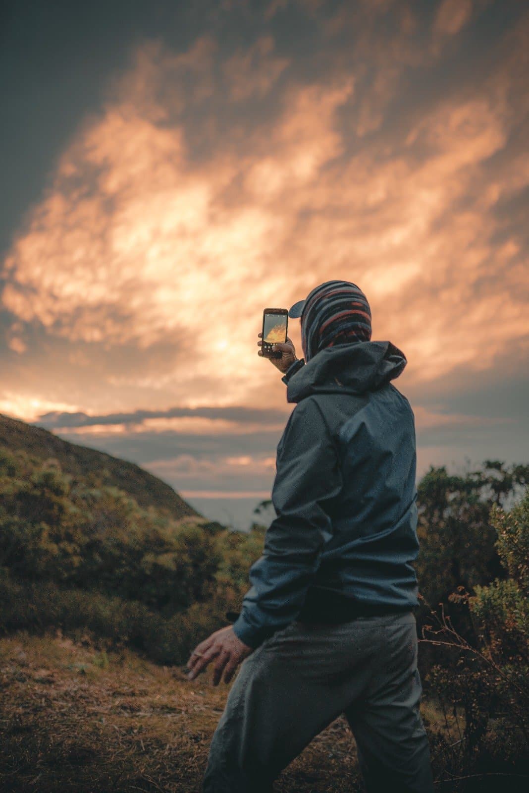 Manche Himmel dulden keinen Aufschub, also bleibt man einfach stehen. Ein Wanderer in Jacke und gestreifter Mütze fotografiert mit dem Smartphone einen dramatischen Sonnenuntergang über dem Kamm der Serra da Mantiqueira beim Serra Fina Trekking