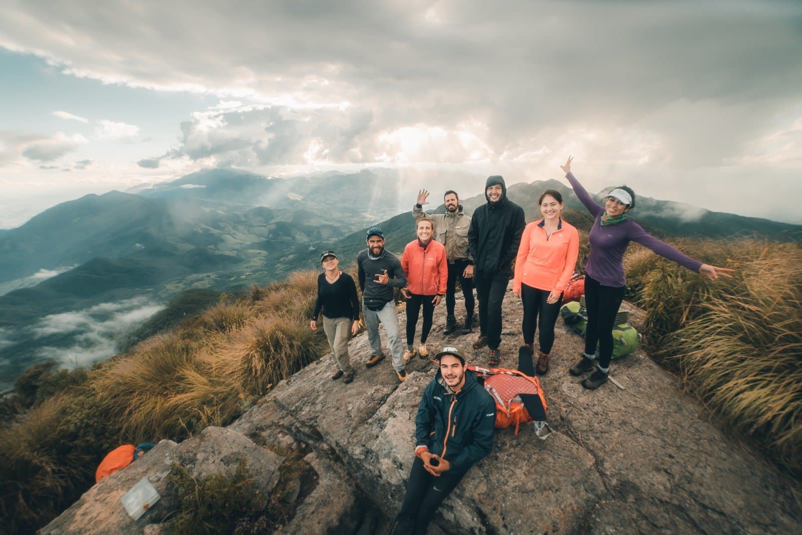 Oben angekommen, und die Wolken machen gleich mit. Acht Wanderer feiern auf einem Felsgipfel beim Serra Fina Trekking, mit dramatischen Wolken und den Bergkämmen der Serra da Mantiqueira im Hintergrund