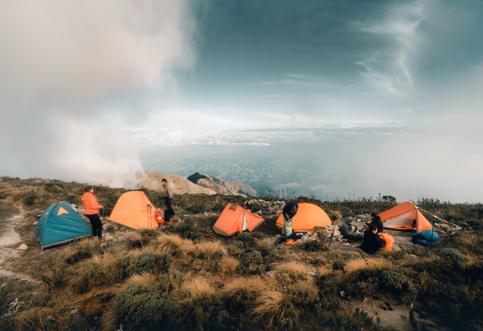 Zelte stehen, Wolken ziehen auf, die Stimmung bleibt gut. Eine Gruppe von Trekkern schlägt orange und blaue Zelte auf einem exponierten Höhenkamm des Serra Fina Trekkings in der Serra da Mantiqueira auf, mit weitem Blick über Täler und dramatischen Gewitterwolken