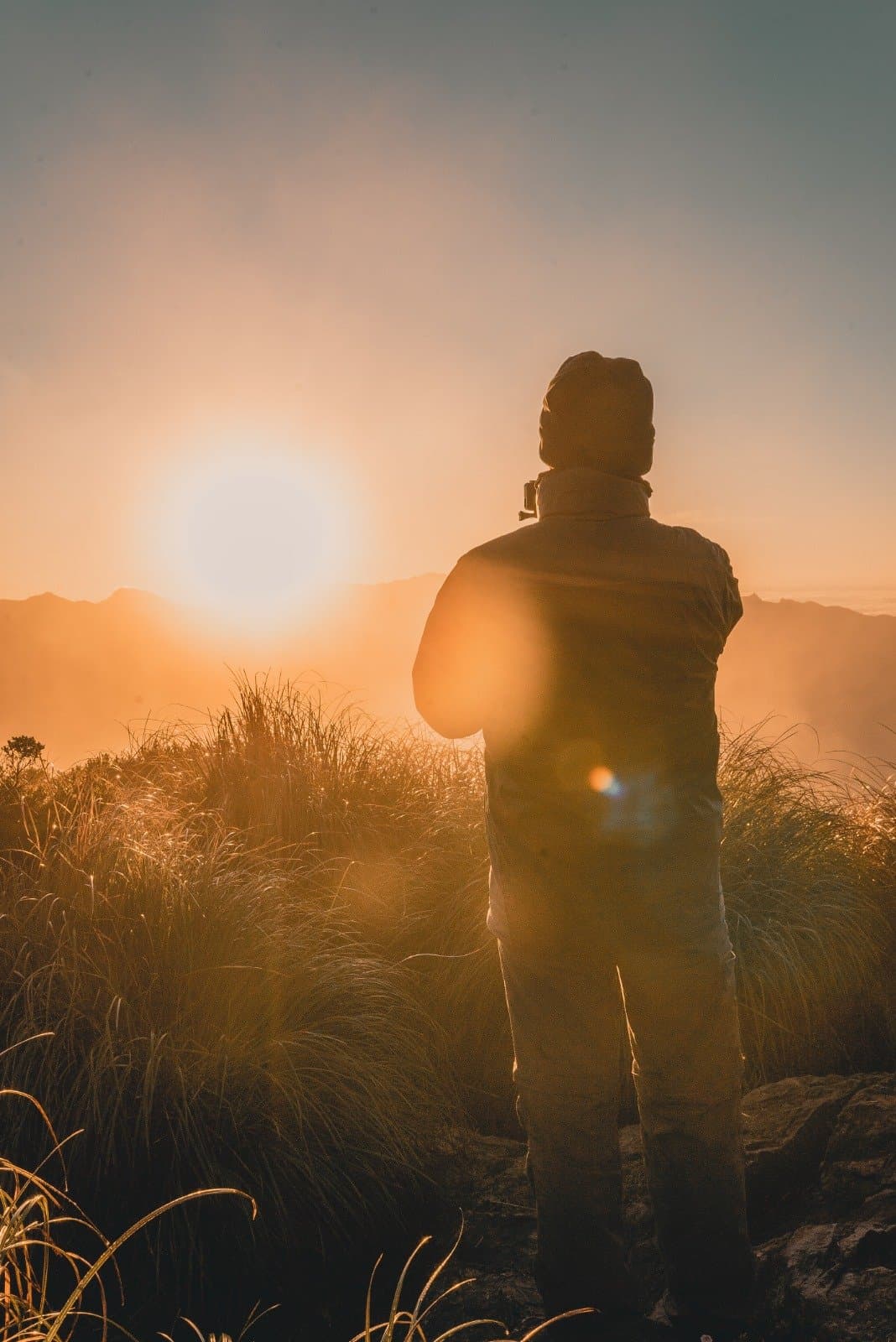 First light over the Mantiqueira — worth every steep step. A hiker's silhouette stands among golden mountain grasses at sunrise during a Serra Fina Trekking expedition, with misty ridges of the Serra da Mantiqueira glowing orange in the background