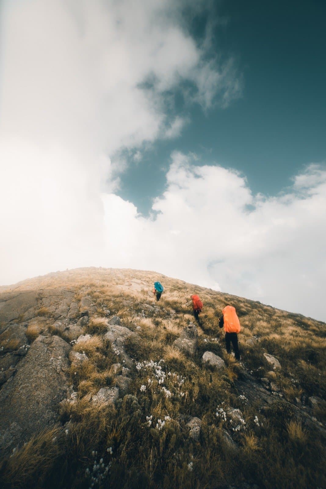 Oben wartet die Aussicht, unten bleibt die Entscheidung, aufzubrechen. Drei Wanderer mit bunten Rucksäcken erklimmen einen steilen Felsgrat beim Serra Fina Trekking in der Serra da Mantiqueira unter einem dramatischen Wolkenhimmel