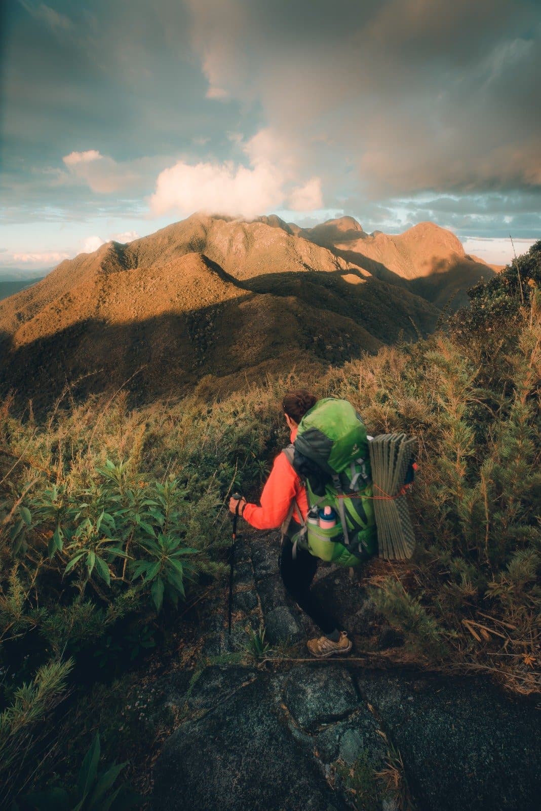 The ridge opens up — and the Mantiqueira takes your breath away. A backpacker in an orange jacket carrying a full pack pauses on a rocky ridge during Serra Fina Trekking, with golden-lit mountain summits of the Serra da Mantiqueira stretching under a dramatic cloudy sky