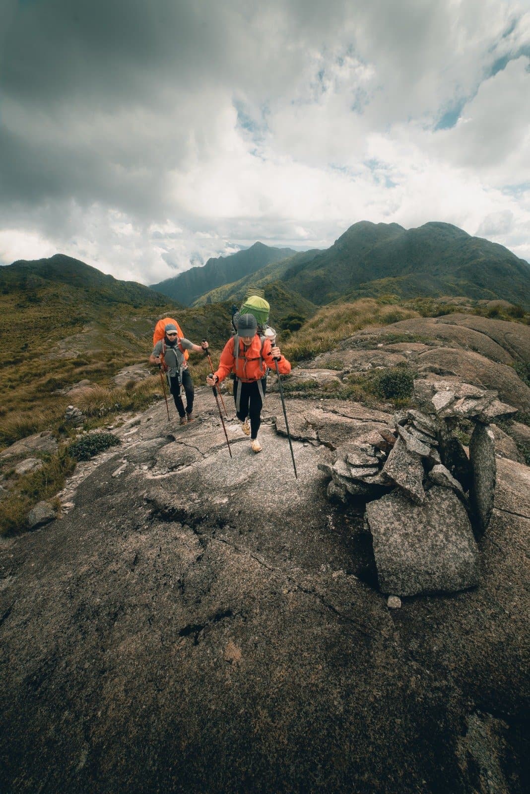 Granit unter den Stiefeln, Wolken im Nacken, genau so soll Trekking sein. Zwei Trekker in orangefarbenen Jacken mit Trekkingstöcken überqueren eine Granitfelsplatte beim Serra Fina Trekking, im Hintergrund die grünen Bergrücken der Serra da Mantiqueira unter einem dramatischen Wolkenhimmel