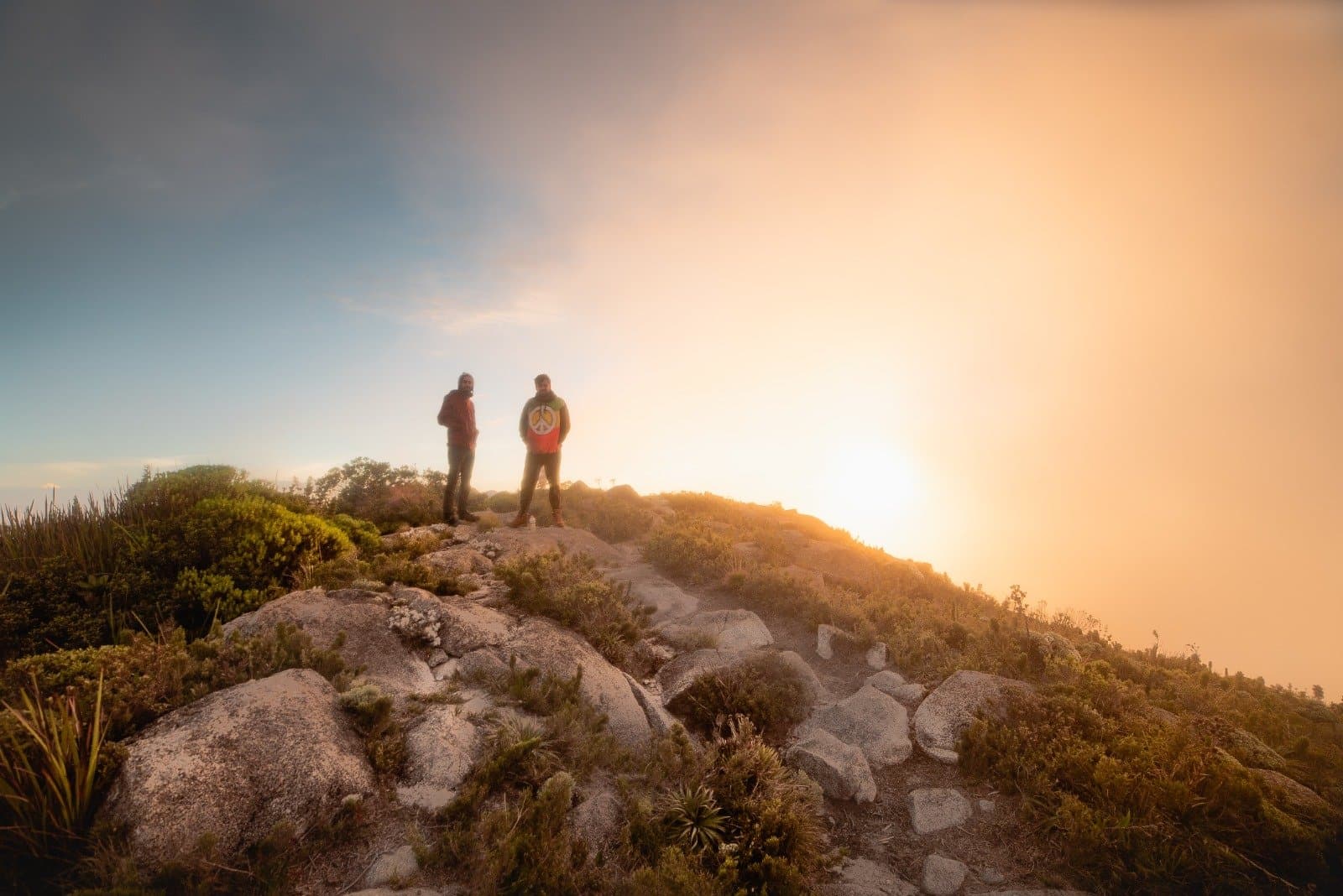 Früh aufstehen nervt. Dieser Moment macht es wieder gut. Zwei Wanderer als Silhouette vor einem strahlend goldenen Sonnenaufgang auf dem felsigen Gipfel des Serra Fina Trekkings in der Serra da Mantiqueira, umgeben von Hochlagenvegetation des Atlantischen Regenwaldes.