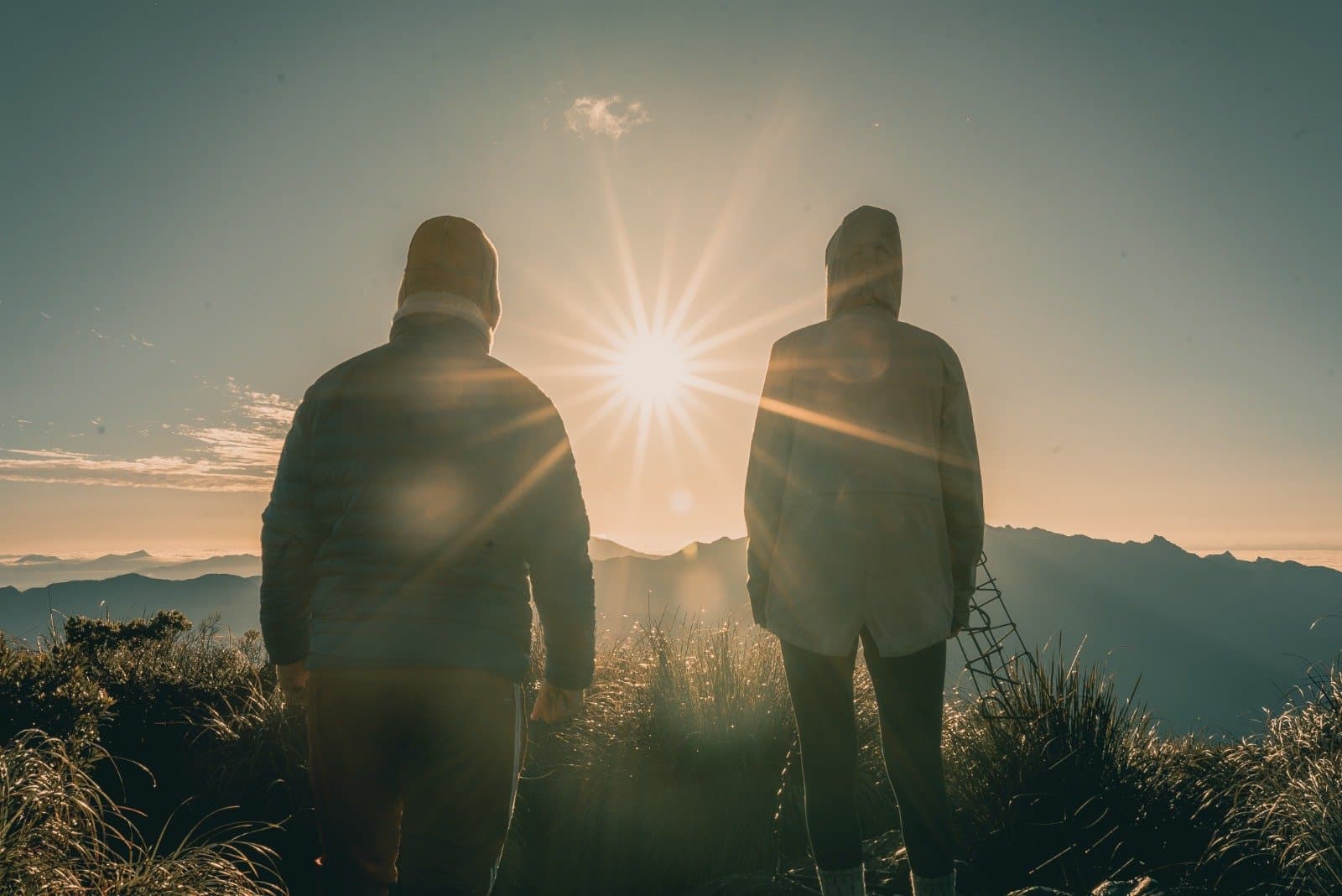 Der Berg redet, die zwei hören einfach zu. Zwei Wanderer mit Kapuzenjacken stehen als Silhouette gegen einen strahlenden Sonnenaufgang auf einem Gipfel des Serra Fina Trekkings, mit den Bergketten der Mantiqueira im Hintergrund