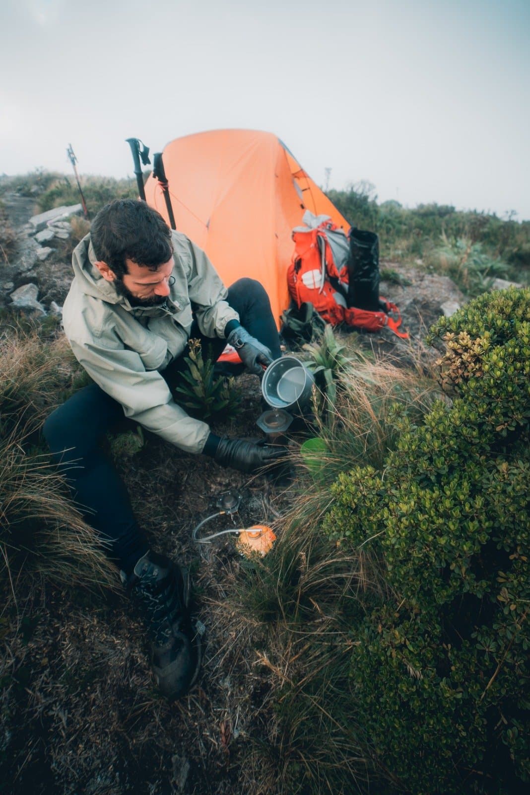 Warmes Essen, kalte Finger, und das Gefühl, genau am richtigen Ort zu sein. Ein bärtiger Wanderer in Regenjacke gießt Wasser in einen Topf auf einem Campingkocher neben einem orangefarbenen Zelt beim Serra Fina Trekking in der Serra da Mantiqueira