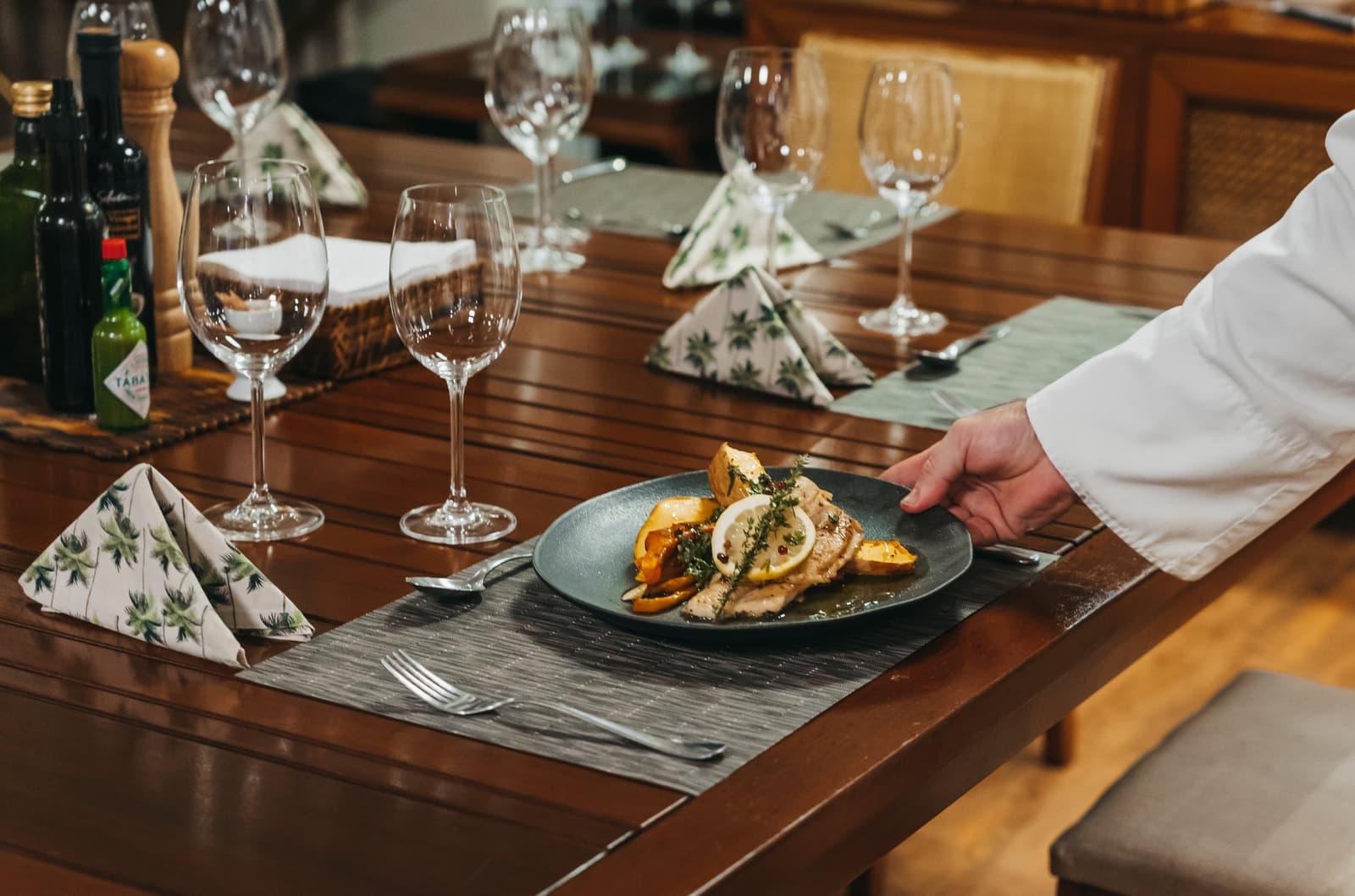 Unhurried, immaculate — dinner served in the heart of the rainforest. A chef in a white jacket placing a dark plate of grilled fish with roasted peppers, lemon and fresh herbs on a formally set wooden dining table aboard the untamed amazon cruise