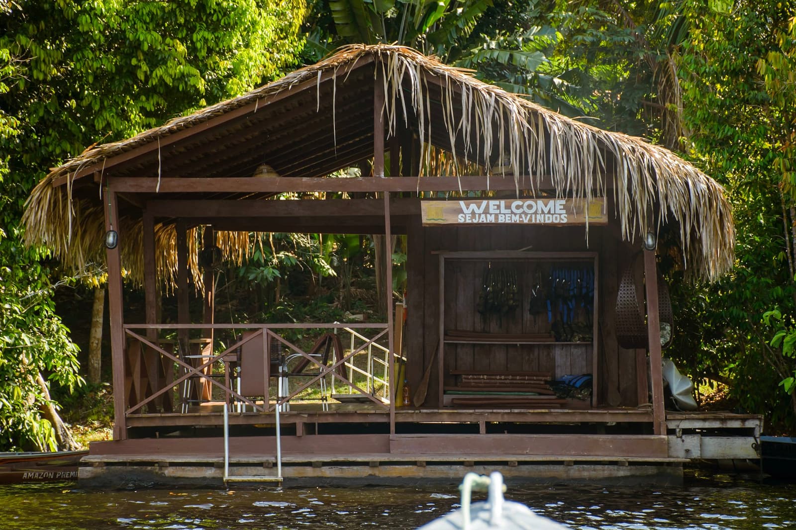 Chegar de barco e ver a placa de boas-vindas, o roteiro começa bem. Um pavilhão de atracação em madeira com telhado de palha e placa 'Welcome / Sejam Bem-Vindos', fotografado da água durante um roteiro pela Amazônia e Pantanal, cercado por vegetação densa