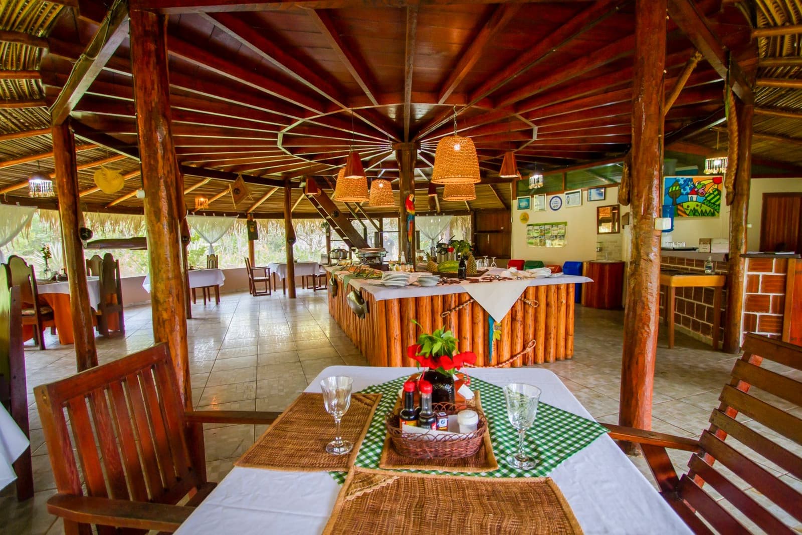 Mesa posta, brisa do cerúleo e o Pantanal logo ali fora. Salão de refeições ao ar livre com teto de palha em uma pousada do Pantanal durante um passeio pela Amazônia e Pantanal, com buffet rústico revestido de toras, luminárias de vime, mesas cobertas com toalhas brancas e taças de vinho, abertas para a vegetação