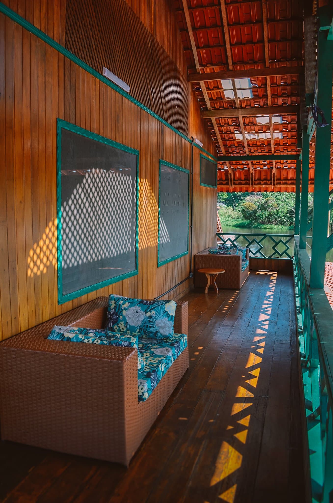 Middaglicht door het gaas, de rivier op een steenworp afstand. Overdekt houten terras van een verblijf tijdens een amazon rainforest ecolodge trip in Brazilië, met rieten stoelen, gebloemde kussens, gaasramen die ruitvormige schaduwen werpen en uitzicht op groene oevervegetatie