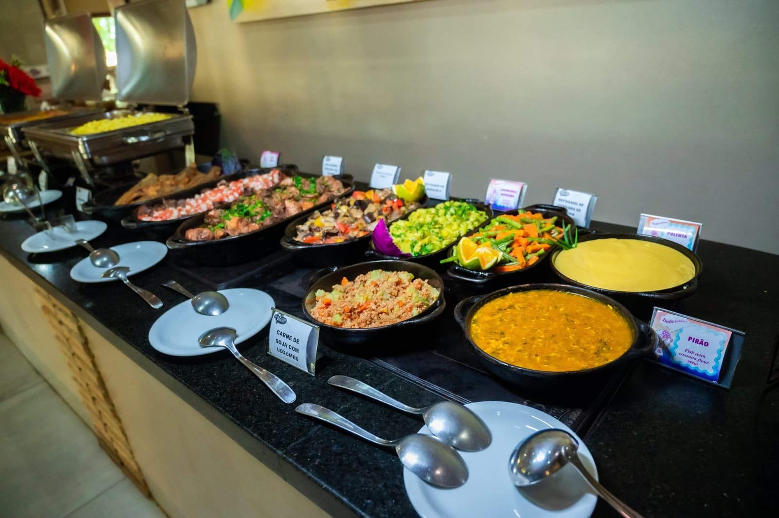 Pirão, polenta, carne de soja, as plaquinhas identificam, o apetite aprova. Mesa de buffet brasileiro em um tour pelo Amazonas e Pantanal, com pratos identificados como pirão, polenta, carne de soja com legumes, legumes refogados com rodelas de laranja e farofa, servidos em panelas de barro preto sobre balcão de granito escuro