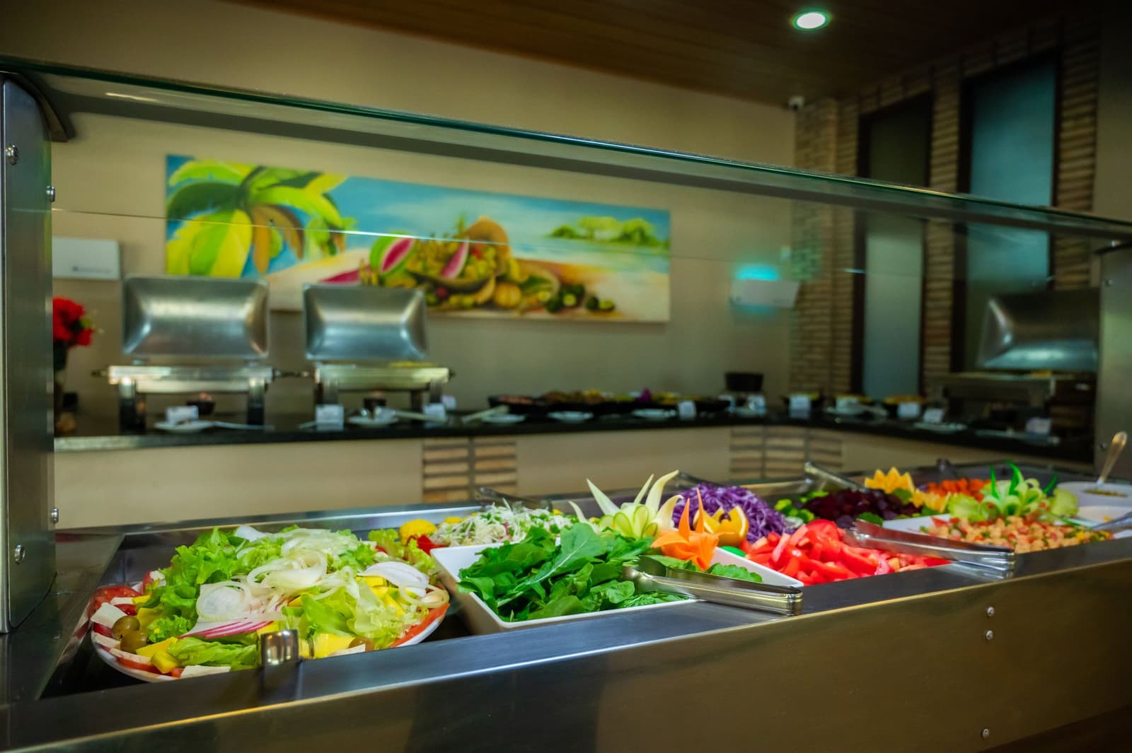 Prato cheio de cor antes de mais um dia cheio de natureza. Balcão de buffet de saladas bem servido em um tour pela Amazônia e Pantanal, com bandejas de alface fresca, espinafre, repolho roxo, tomate fatiado e enfeites de legumes esculpidos sob um vidro protetor