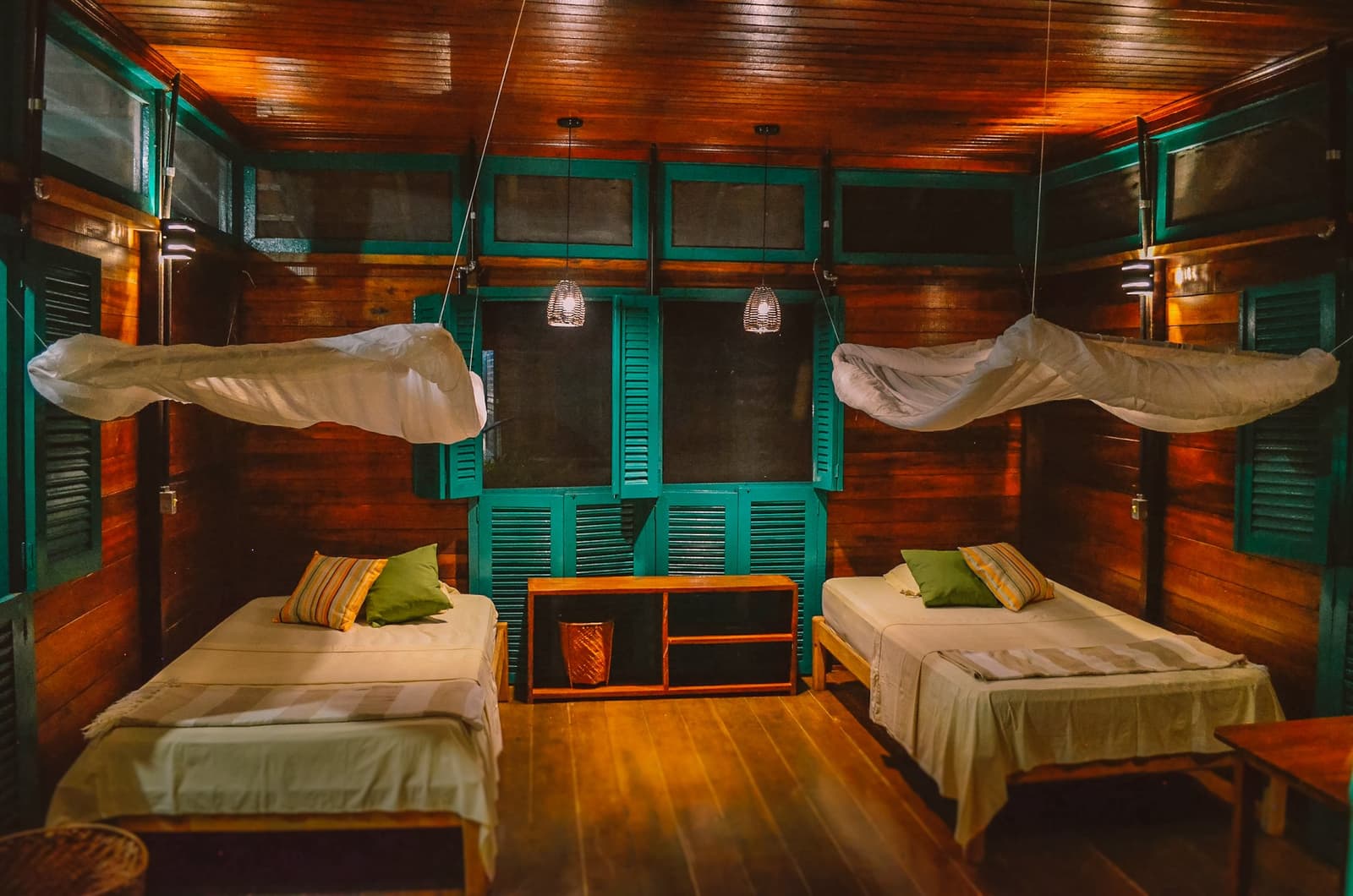 Hout, klamboe en echte rust, meer heb je niet nodig. Tweepersoonskamer in een drijvende Amazone-ecolodge met warme houten wanden en plafond, groenblauw gelambriseerde luiken, klamboes boven elk bed en gevlochten hanglampen die een zacht amberkleurig licht geven