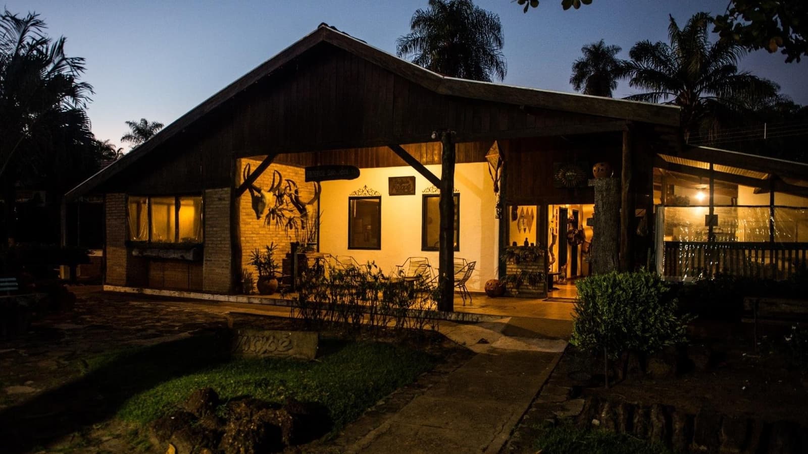 Dinner is almost ready, and the night safari is next. A rustic wooden lodge glowing with warm amber light at dusk on a pantanal bonito tour, with decorative wall mounts, open veranda seating, and palm trees silhouetted against a deep blue twilight sky