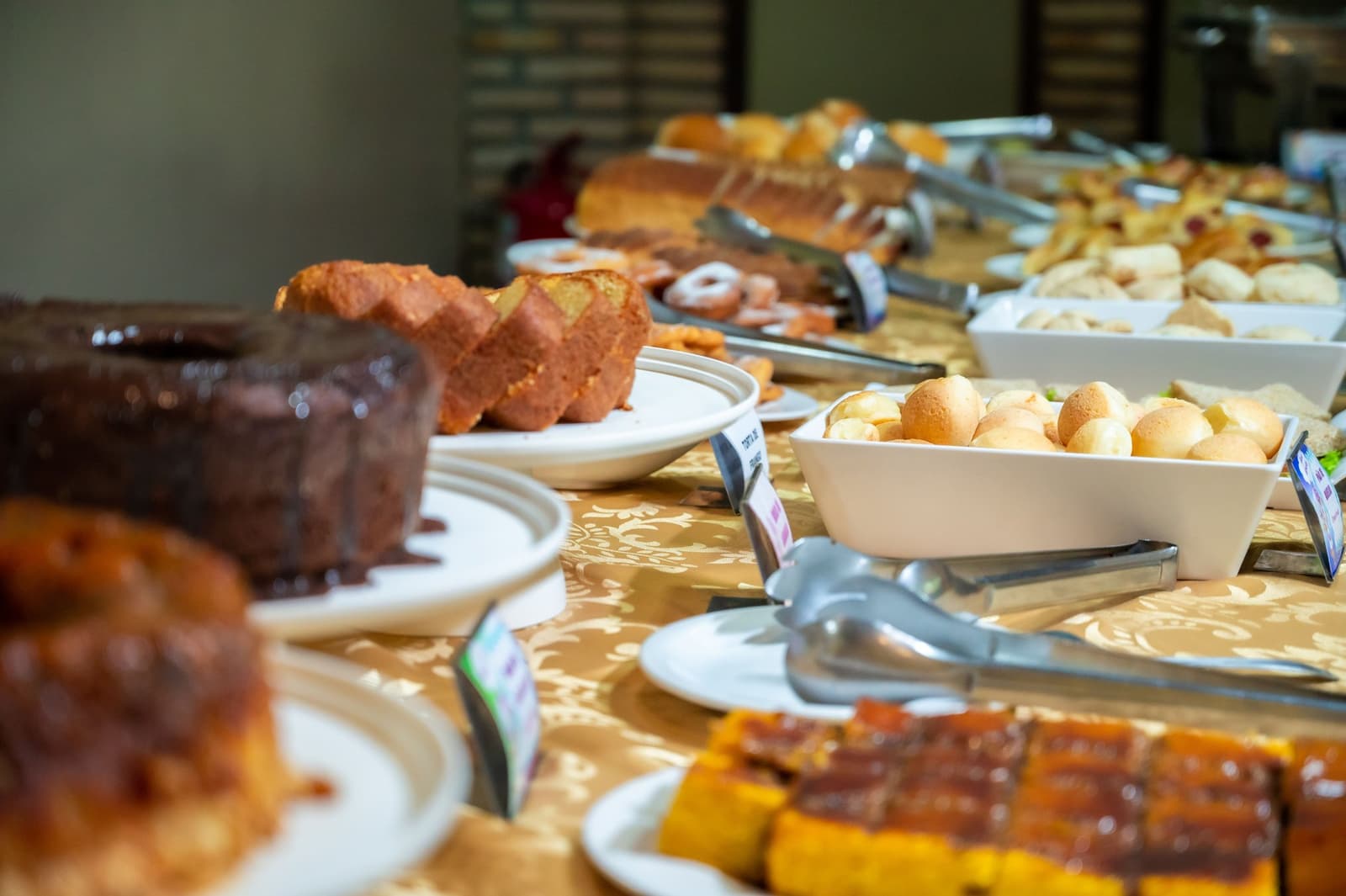 Café da manhã farto antes de jaguar, porque a manhã é longa e boa. Uma longa mesa de café da manhã em um tour pelo Pantanal e Amazônia repleta de fatias de bolo, pão de queijo, pãezinhos doces e quitutes com calda sobre toalha dourada