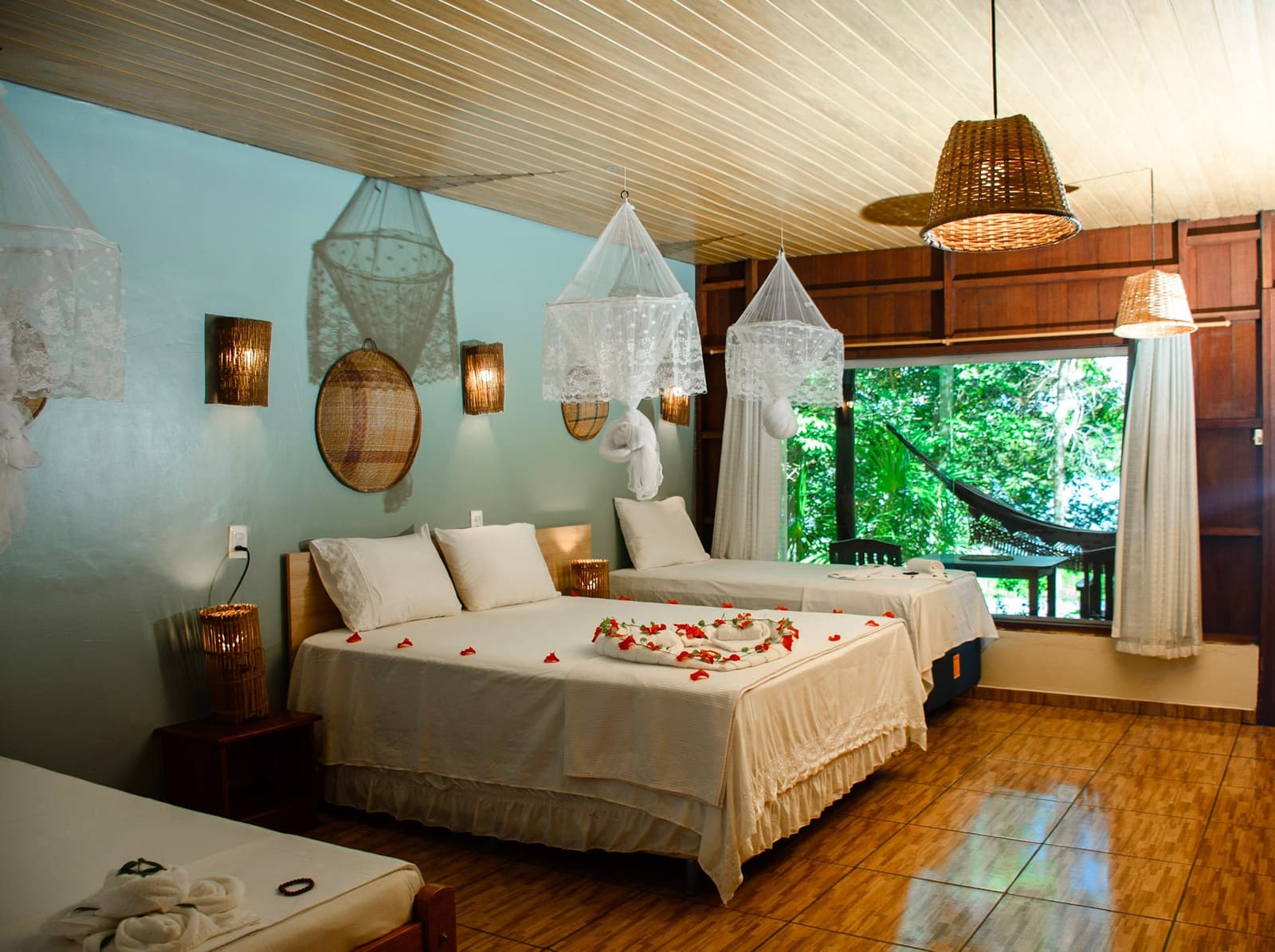 Pétalas na cama, uma rede logo ali pela janela. O roteiro começa bem. Quarto aconchegante de lodge na selva em um roteiro Amazon e Pantanal, com duas camas brancas decoradas com pétalas de flores vermelhas e arte em toalhas, mosquiteiros de renda suspensos no teto, luminárias de vime e uma janela ampla com vista para uma rede e floresta verde