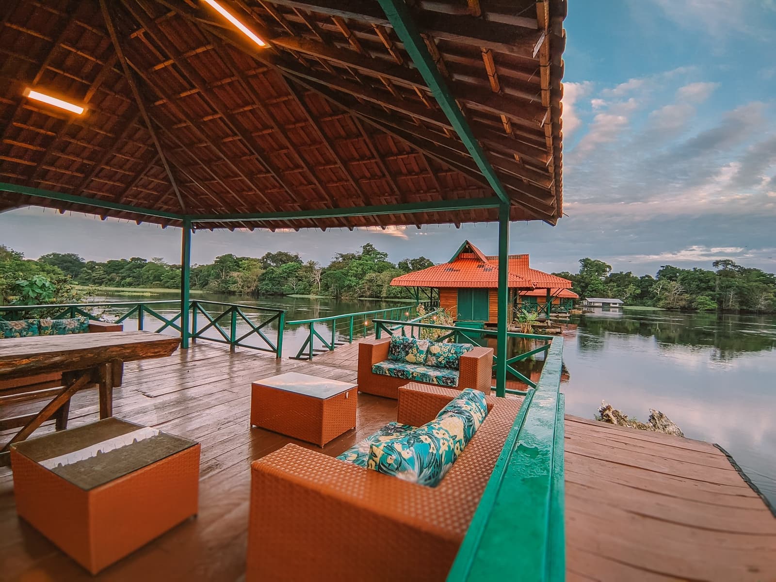 Dit is de lounge, en de Amazone is de achtertuin. Overdekt houten terras van de drijvende Uakari lodge tijdens een Amazon regenwoud ecolodge reis in Brazilië, met rotan banken en bloemetjeskussens met uitzicht op een kalm rivierkanaal en beboste oevers bij schemering