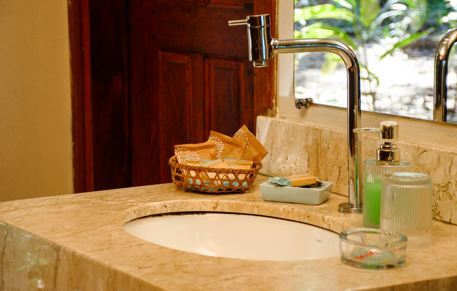 A cestinha de vime com sabonetes é um desses detalhes simples que fazem toda a diferença. Cuba de banheiro com tampo de pedra em uma pousada de roteiro amazônia e pantanal, com torneira cromada, cestinha de vime com amenidades e produtos verdes ao lado de janela com vista para vegetação tropical