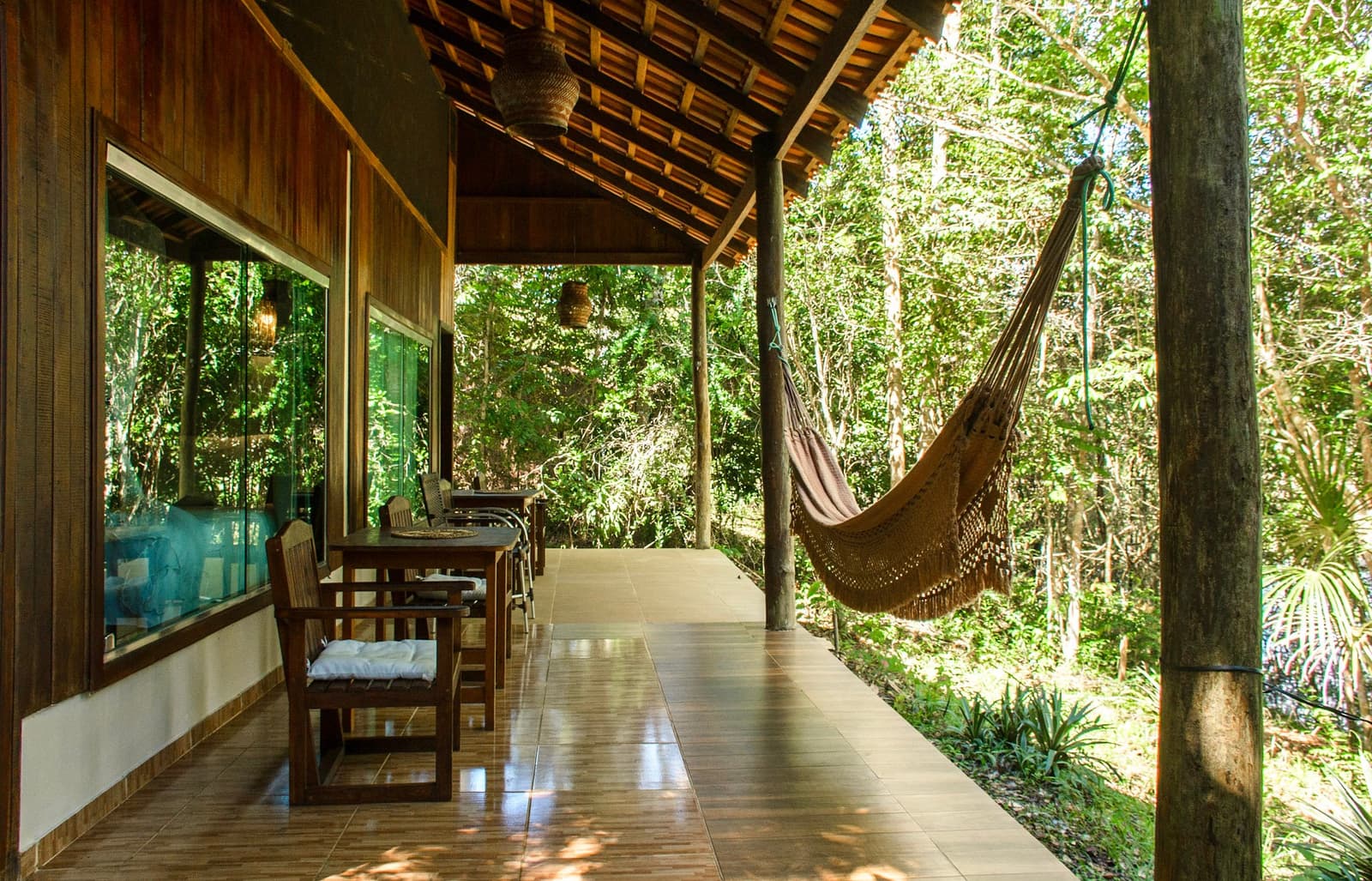 Rede na altura da copa das árvores. Alguns convites não precisam de resposta. Varanda coberta de uma pousada na selva amazônica com uma rede de algodão com franjas pendurada entre pilares de madeira, mesa de jantar e cadeiras de teca, luminárias de vime e floresta densa logo além da borda aberta