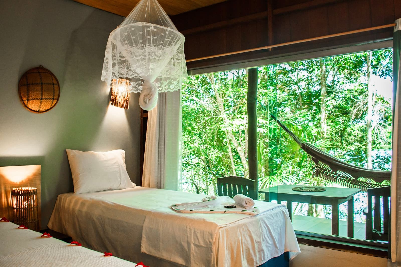 Rede, lençol branco e mata fechada do outro lado do vidro. Quarto duplo em lodge de selva amazônica com camas de linho branco sob um dossel de mosquiteiro de renda, arandela de parede aconchegante, decoração de cesta trançada e janela ampla com vista para a copa verde da floresta e uma rede na varanda