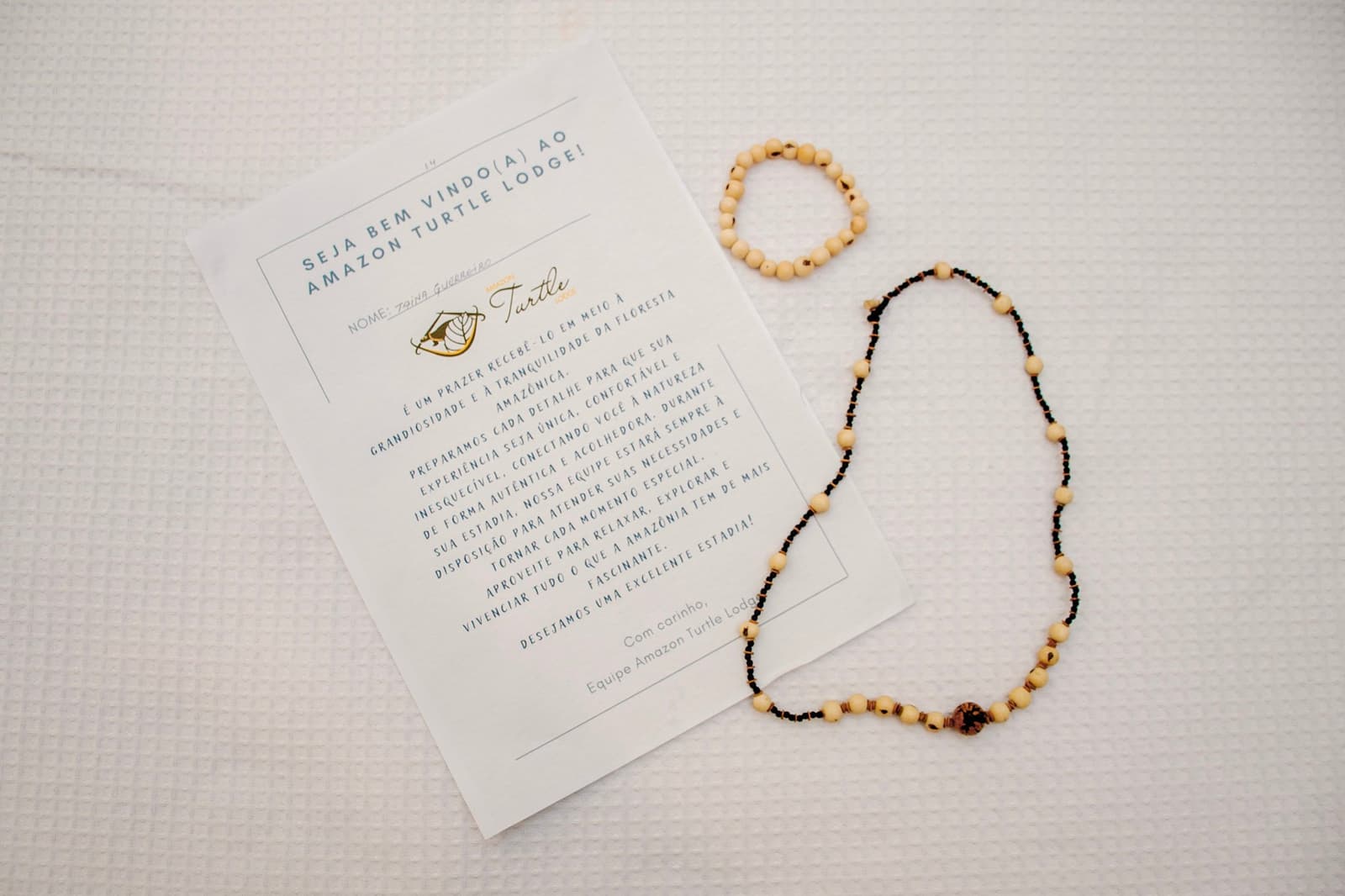 Sementes trabalhadas à mão e um bilhete com o seu nome. Eles estavam te esperando. Cartão de boas-vindas personalizado de um lodge na Amazônia ao lado de um colar e pulseira artesanais de sementes sobre colcha branca de algodão