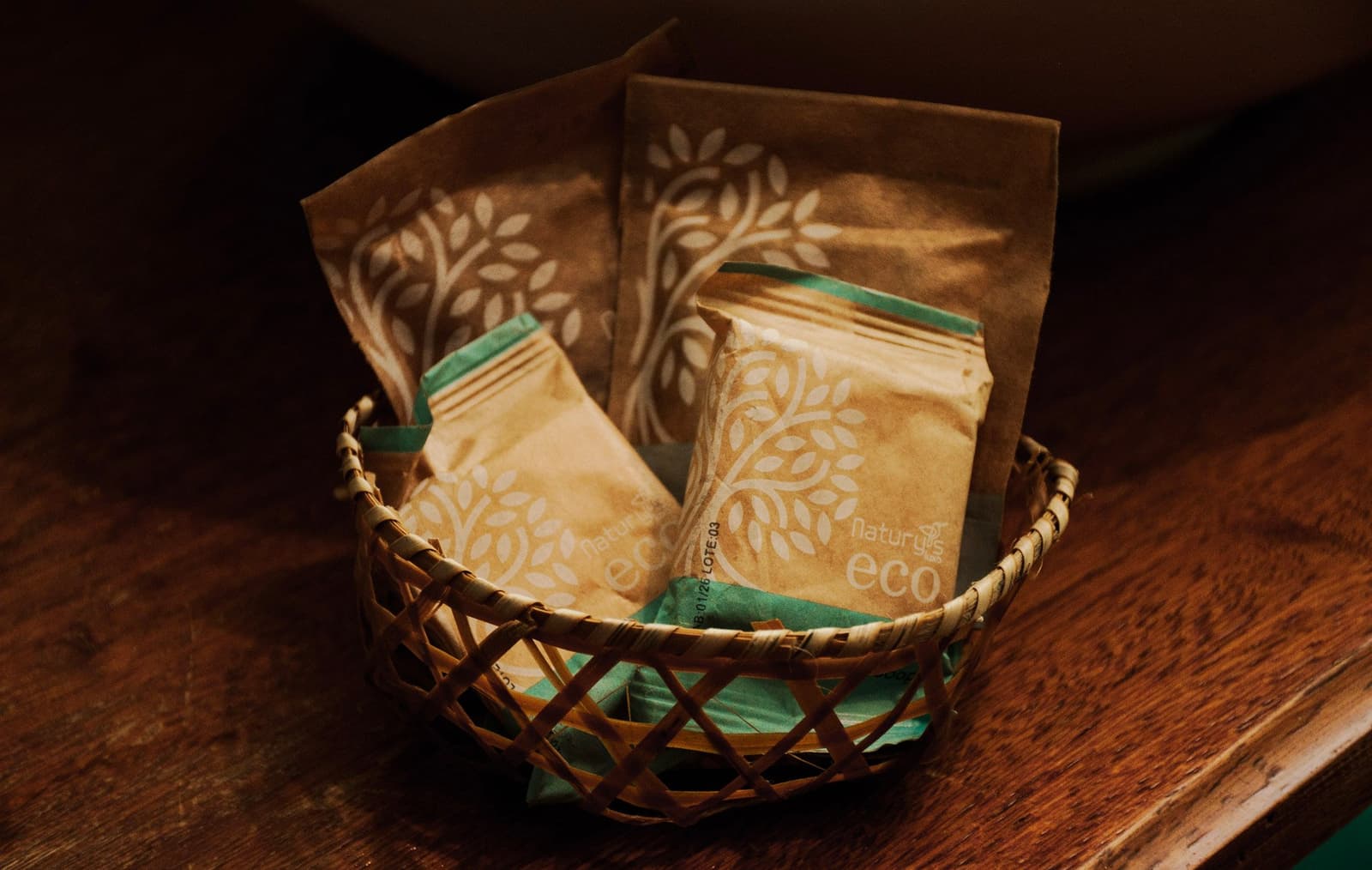 Sem plástico, sem desculpa. O cuidado com a floresta começa nos detalhes do quarto. Quatro sachês de produtos Natury Eco em papel kraft com estampa de folhas, dispostos em uma cestinha trançada sobre superfície de madeira, como amenidades ecológicas de lodge em tour Amazônia e Pantanal