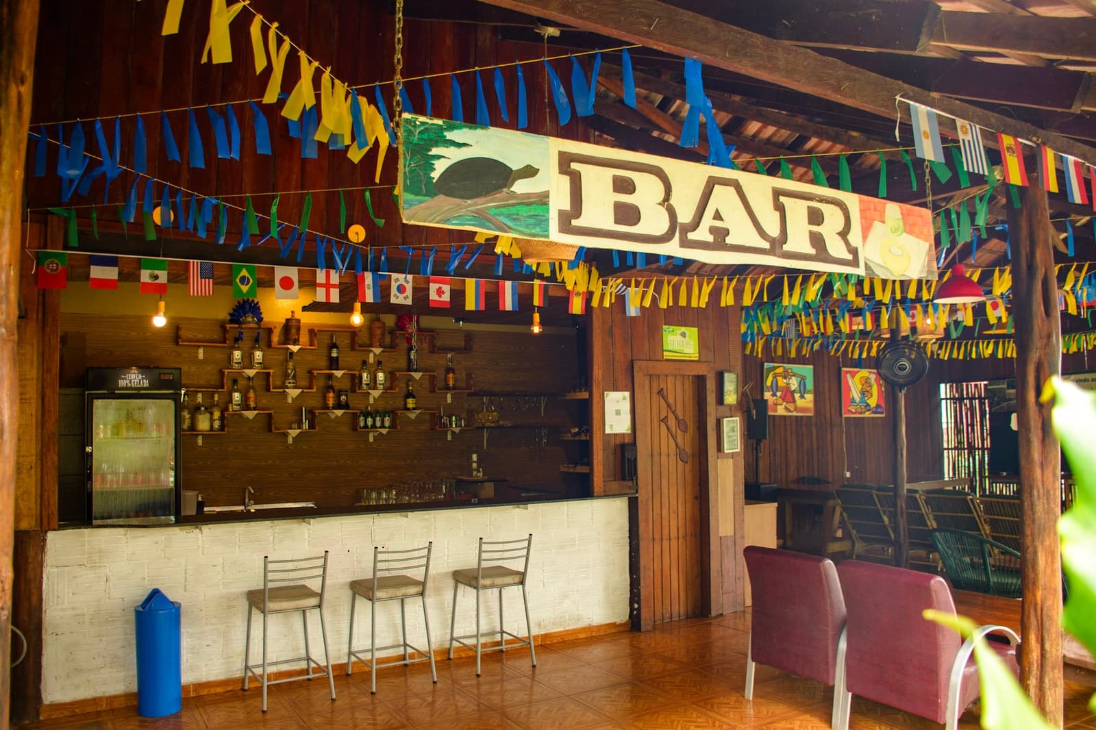 Bandeiras de dezenas de países, uma tartaruga pintada à mão e uma geladeira gelada. O bar faz jus à chegada. Bar rústico ao ar livre de uma lodge na selva durante um roteiro pela Amazônia e Pantanal, decorado com bandeiras internacionais, bandeirolas azuis e amarelas e uma placa pintada à mão com uma tartaruga acima da palavra BAR, com garrafas em prateleiras de madeira e banquetas ao longo do balcão