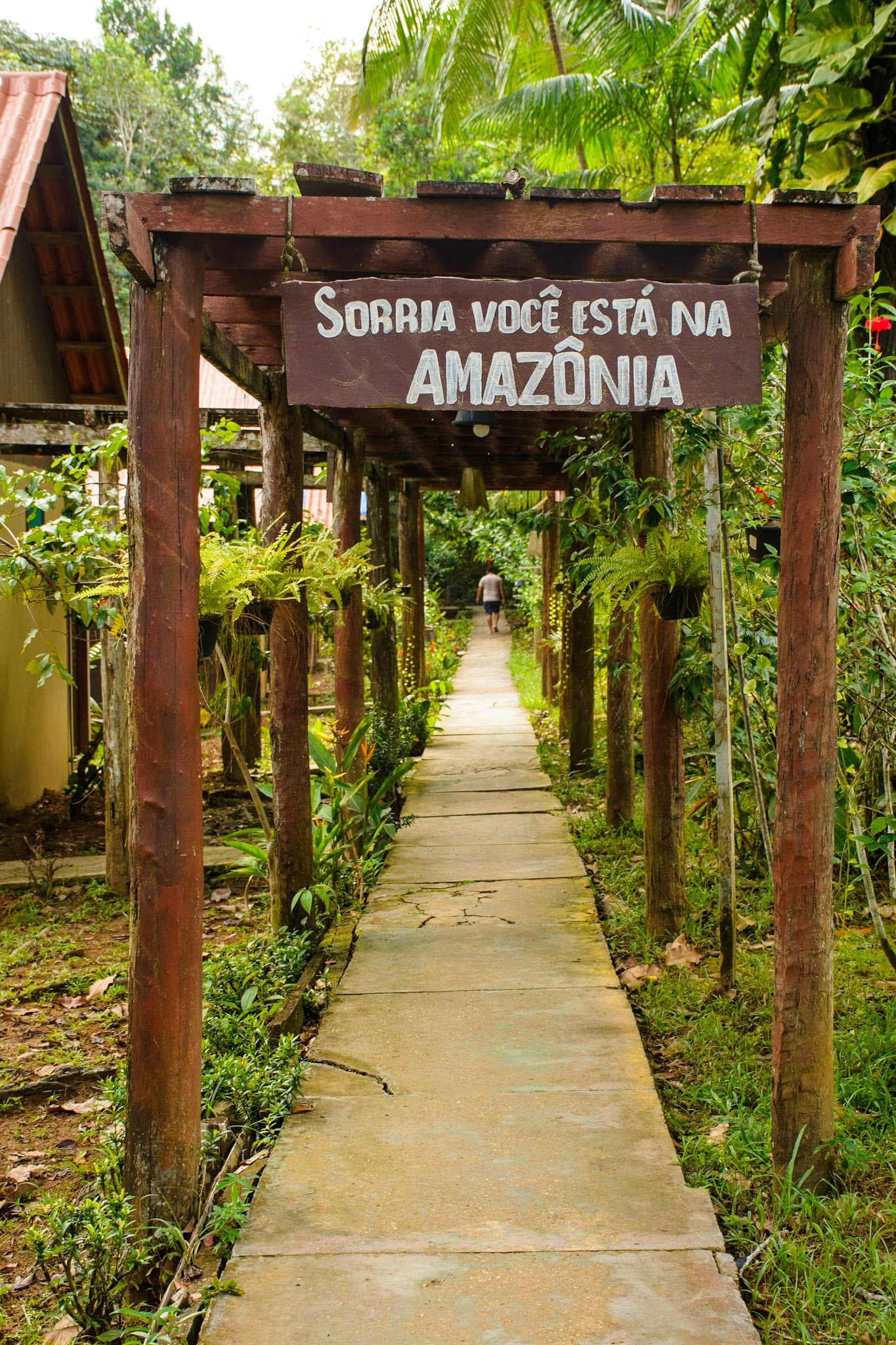 A floresta pede pouco: só um sorriso na chegada. Uma pérgola de madeira rústica na entrada de um lodge na Amazônia exibe uma placa com os dizeres 'Sorria você está na Amazônia', com um caminho de concreto ladeado de vegetação densa e um hóspede caminhando ao fundo.