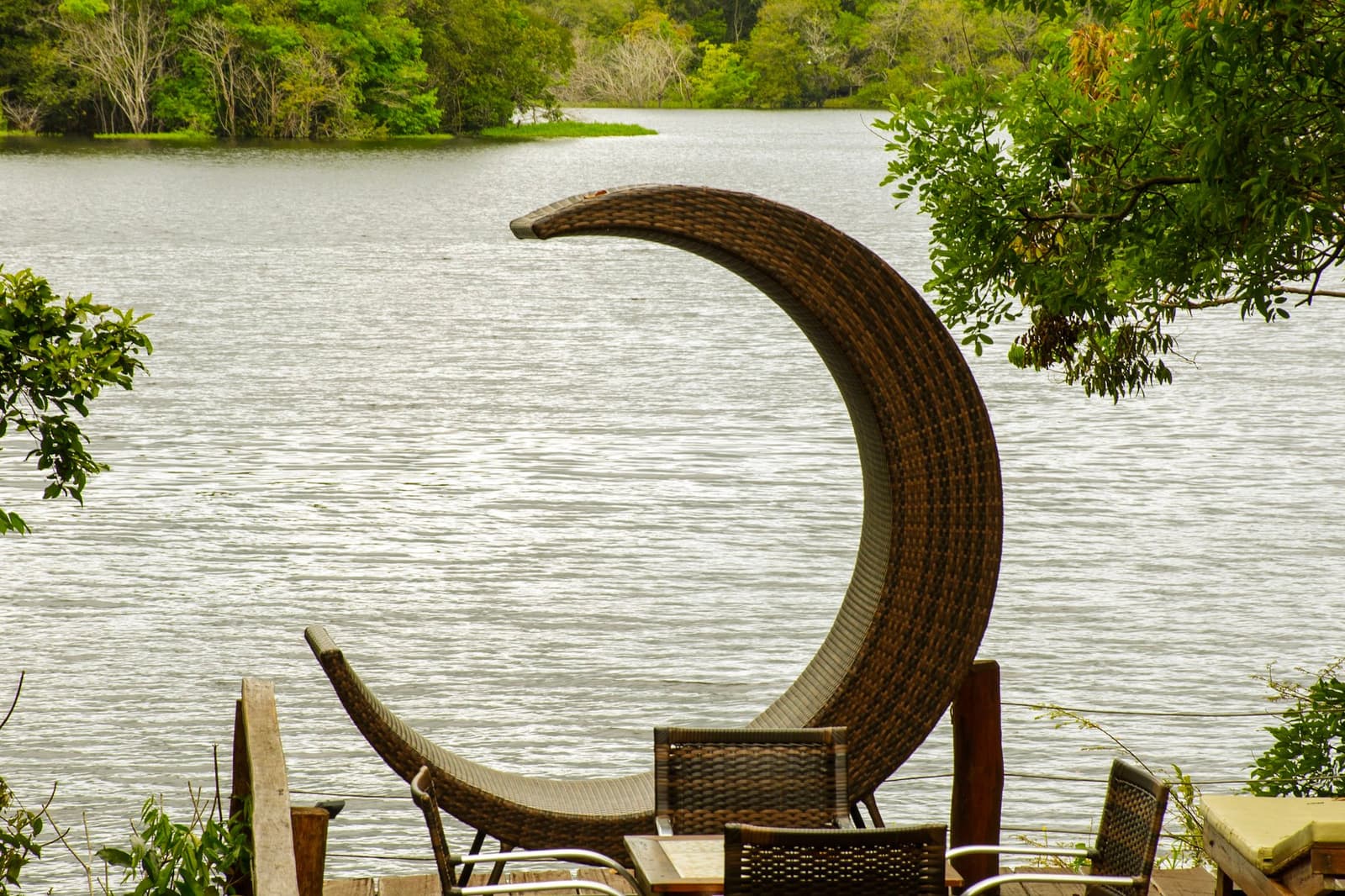 Essa poltrona foi criada por alguém que nunca quis sair da beira do rio. Uma poltrona de vime em formato de meia-lua sobre um deck de madeira com vista para um rio tranquilo ladeado por mata densa, em uma pousada de tour Amazônia e Pantanal