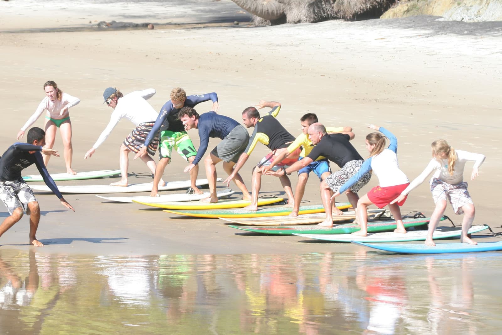 Todo mundo treme na primeira tentativa. Esse é exatamente o começo. Photo: Picasa Um grupo de cerca de dez alunos pratica a posição de pop-up em pranchas de surf durante um exercício em areia seca na escola de surf em Itacaré, enquanto um instrutor local demonstra a postura agachada na frente da fila