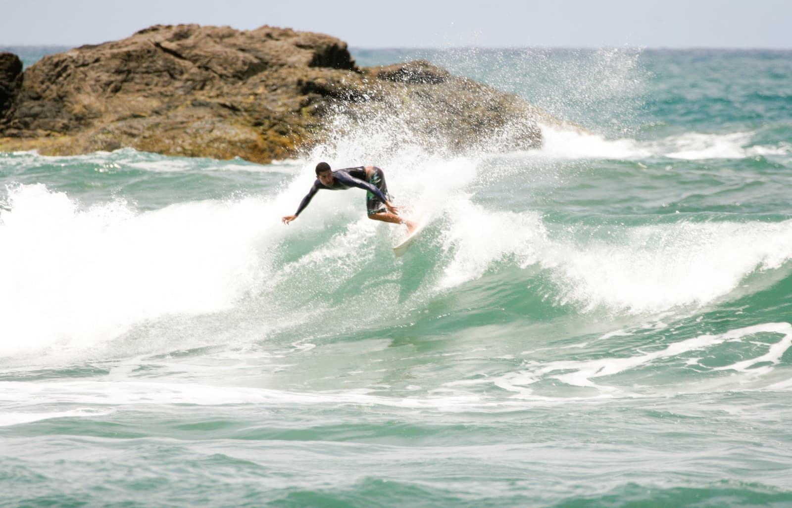 No pico, na hora certa, com a onda escolhida a dedo. Photo: unknown Um surfista com lycra de manga longa executa um cutback preciso em uma onda verde durante uma aula na escola de surf em Itacaré, com uma grande pedra e espuma ao fundo