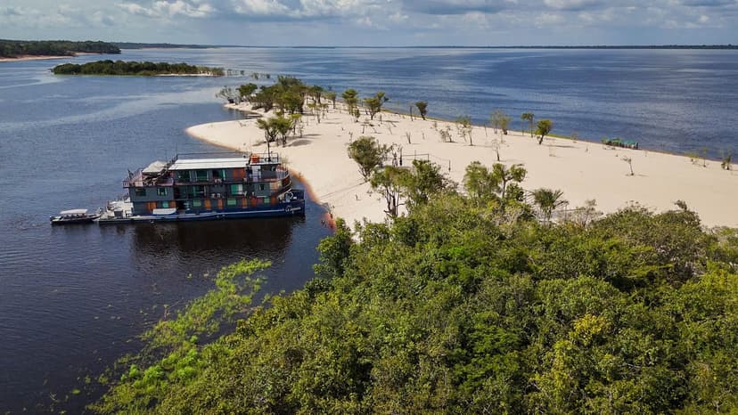 Aerial view of the La Jangada amazon cruise vessel moored beside a white sand beach on a dark-water river, surrounded by dense rainforest canopy and a wide river confluence under a partly cloudy sky. Aerial view of the La Jangada amazon cruise vessel moored beside a white sand beach on a dark-water river, surrounded by dense rainforest canopy and a wide river confluence under a partly cloudy sky.
