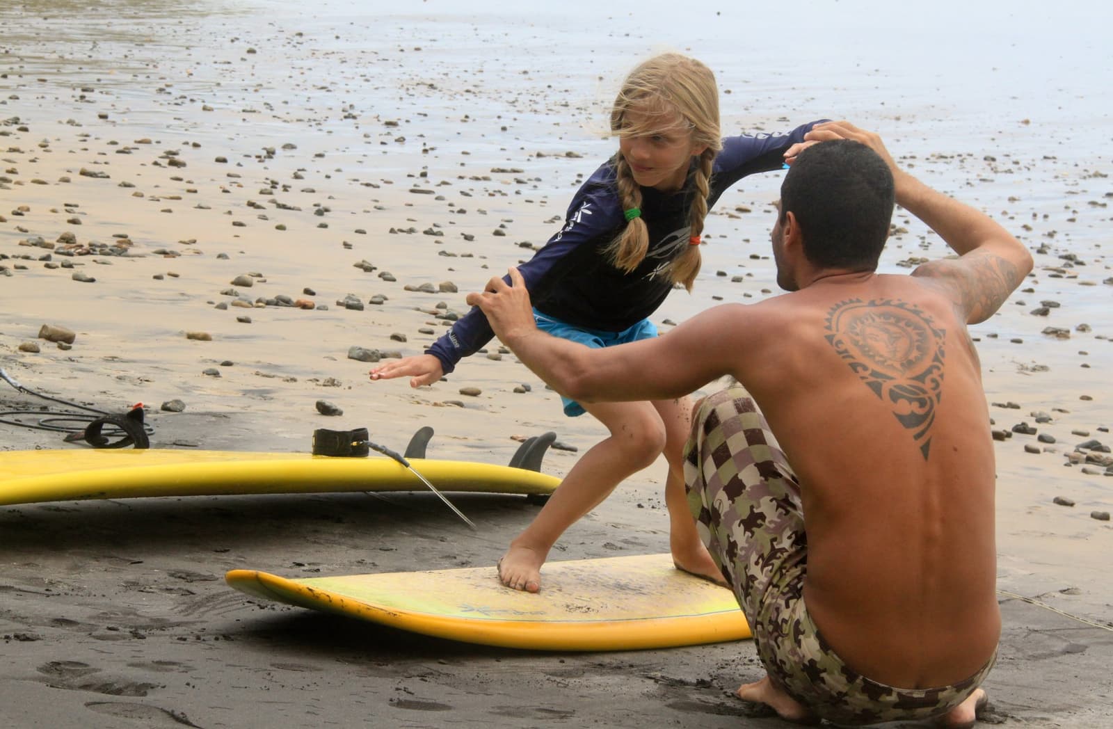 No areia primeiro, na onda depois. É assim que funciona. Photo: Picasa Um instrutor de surf orienta uma jovem que pratica a posição de pop-up numa prancha amarela sobre a areia escura da escola de surf em Itacaré, com planícies rochosas visíveis ao fundo.