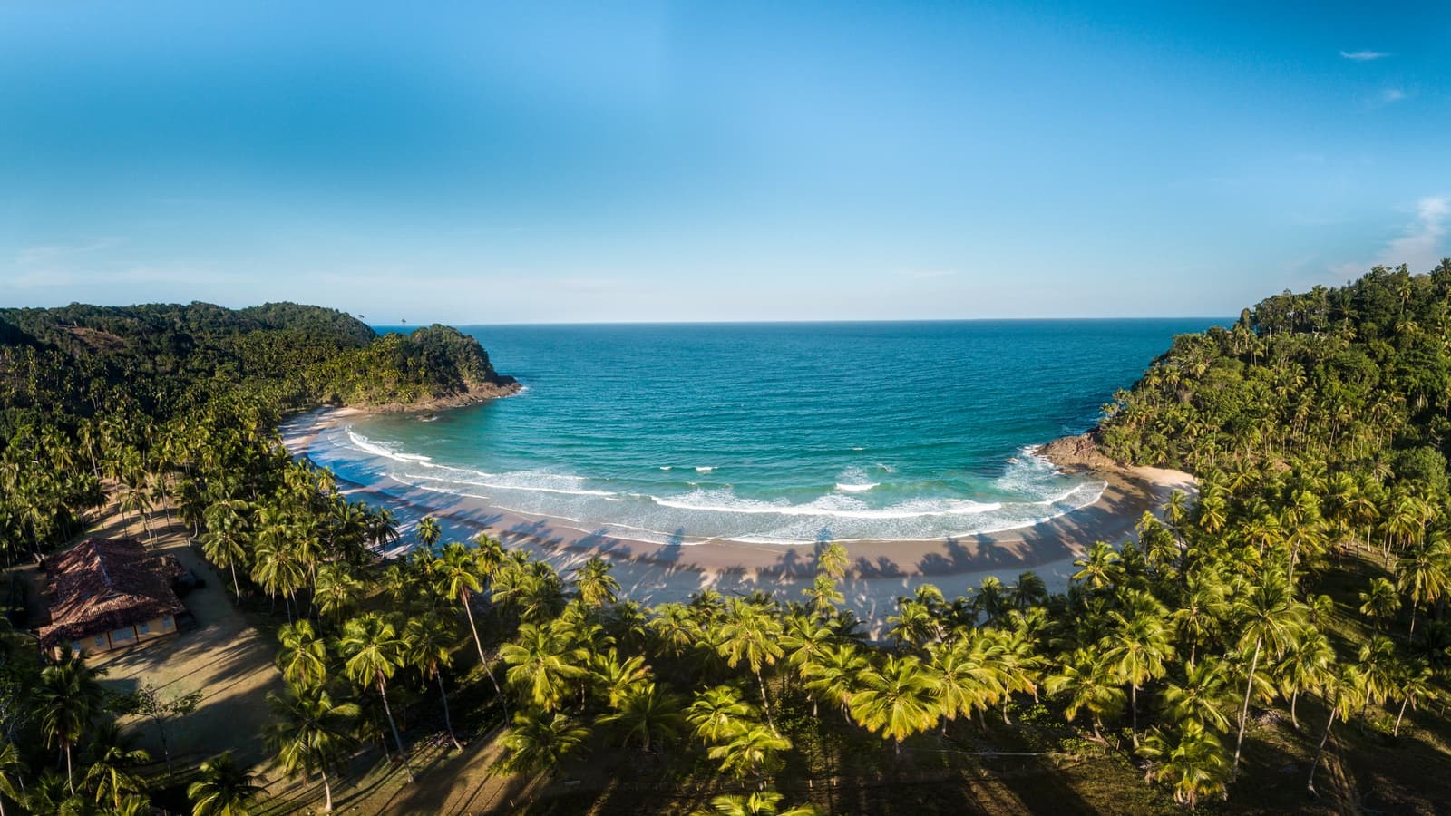 Uma baía tão bem formada que parece ter sido planejada assim. Vista aérea de uma praia em meia-lua cercada por coqueirais densos e Mata Atlântica, com ondas turquesa chegando à areia dourada próximo à costa de Itacaré