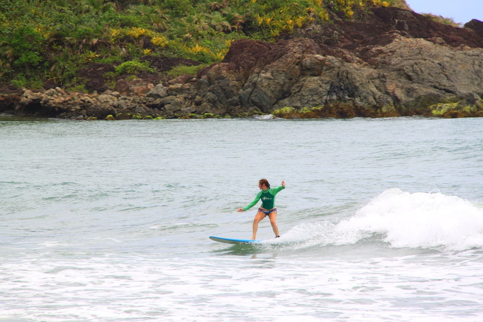 Braços abertos, peso na frente, e de repente tudo faz sentido. Photo: Picasa Uma mulher com lycra verde surfa uma onda verde numa prancha azul de longboard durante uma aula na escola de surf em Itacaré, com um costão rochoso e vegetado ao fundo.