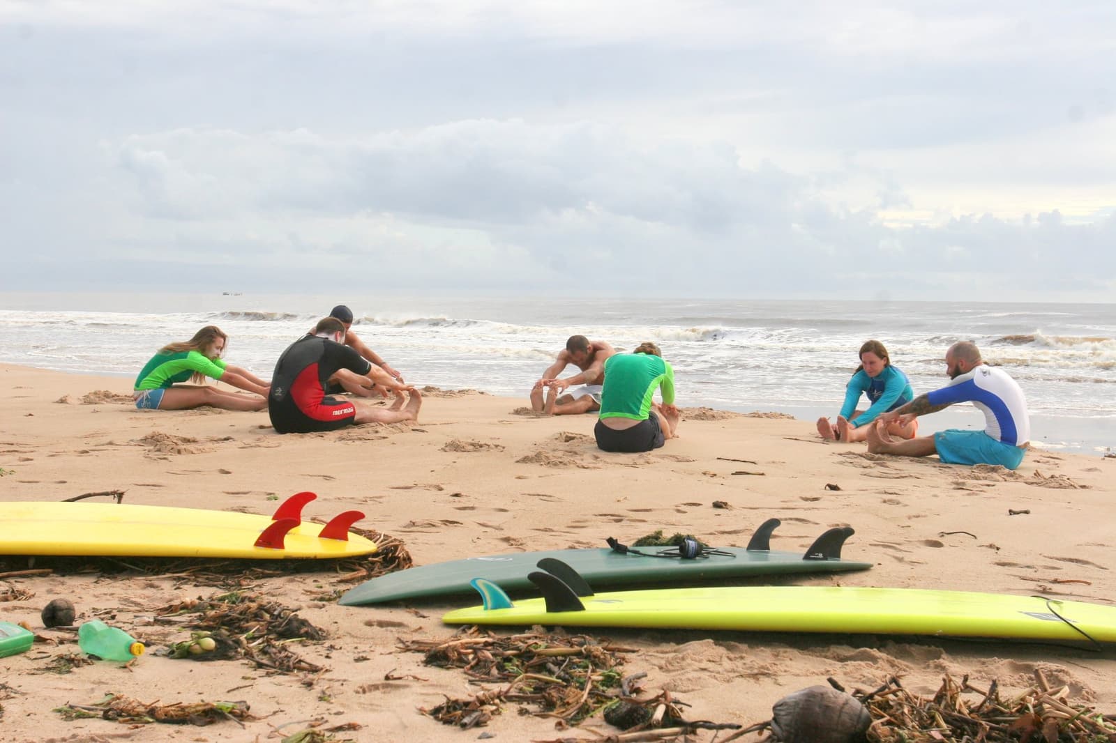 Aquecimento a sério: as costas agradecem antes mesmo de entrar no mar. Photo: Picasa Seis alunos da escola de surf em Itacaré sentados em duplas na areia fazendo alongamento antes da aula, com pranchas no primeiro plano e ondas quebrando ao fundo