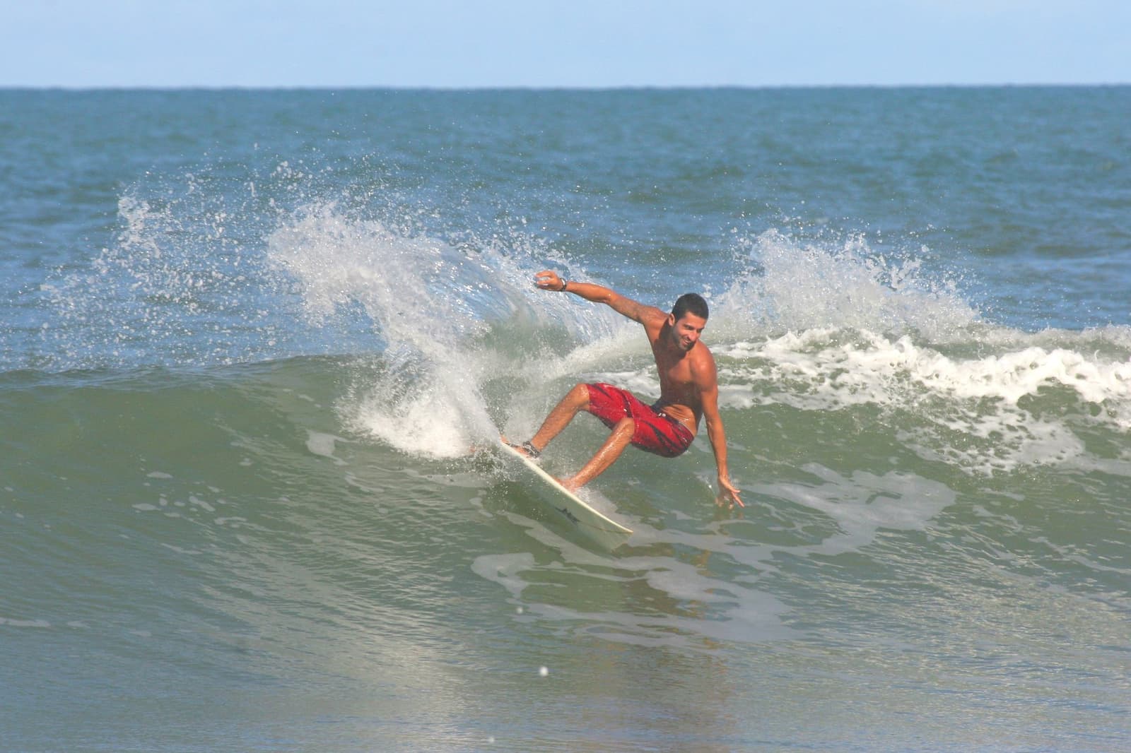 Quatro dias de aula, e o mar já começa a responder. Photo: Picasa Um surfista de bermuda vermelha executa um cutback preciso numa onda verde durante uma aula da escola de surf em Itacaré, braços abertos para equilibrar enquanto o spray branco estoura atrás dele