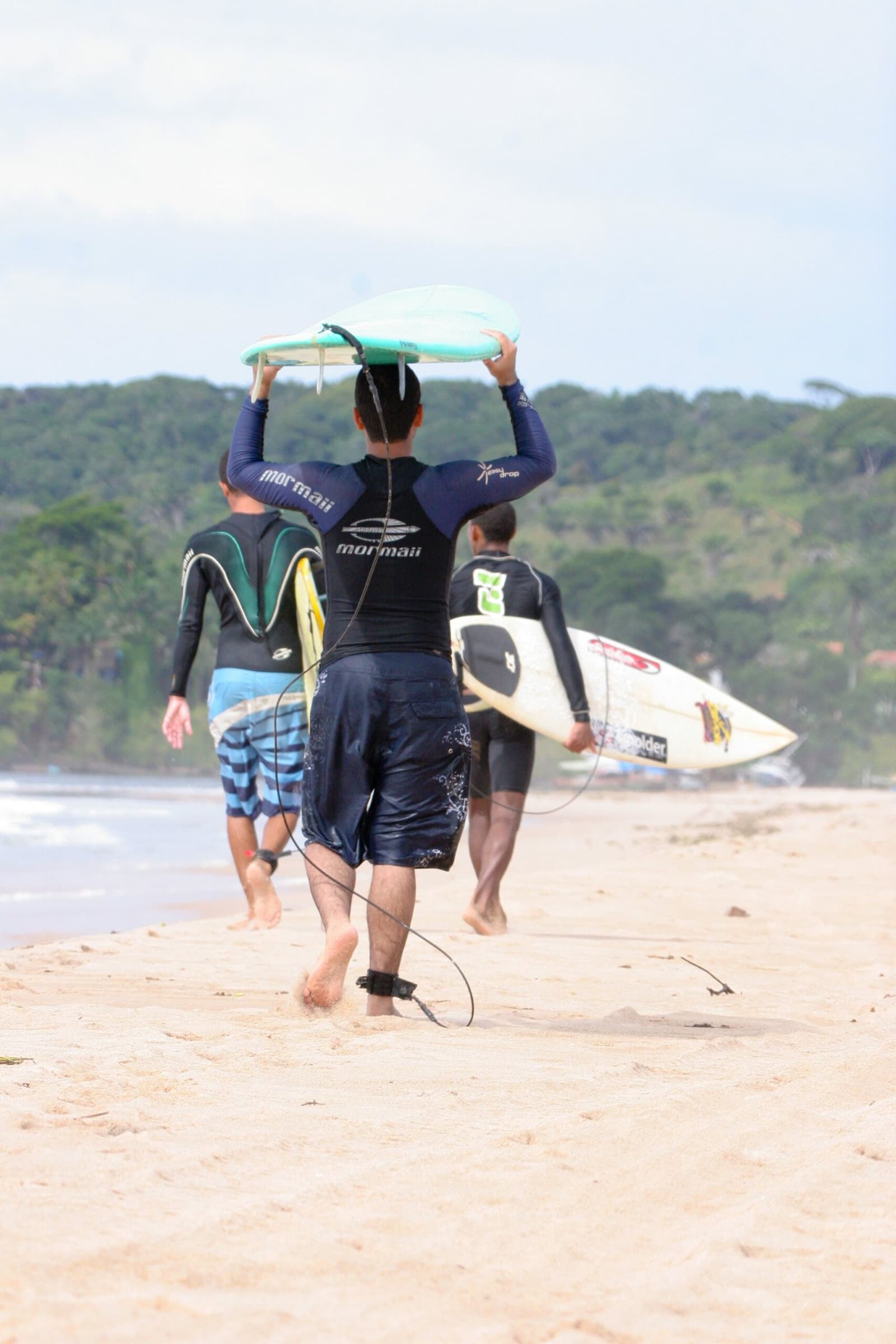 Pranchas no alto, leash no tornozelo, o mar não espera. Photo: unknown Três surfistas de roupa de neoprene e shorts caminhando em direção ao mar numa aula de surf em Itacaré, um com uma prancha turquesa erguida acima da cabeça, outro segurando um shortboard branco, mata atlântica densa ao fundo
