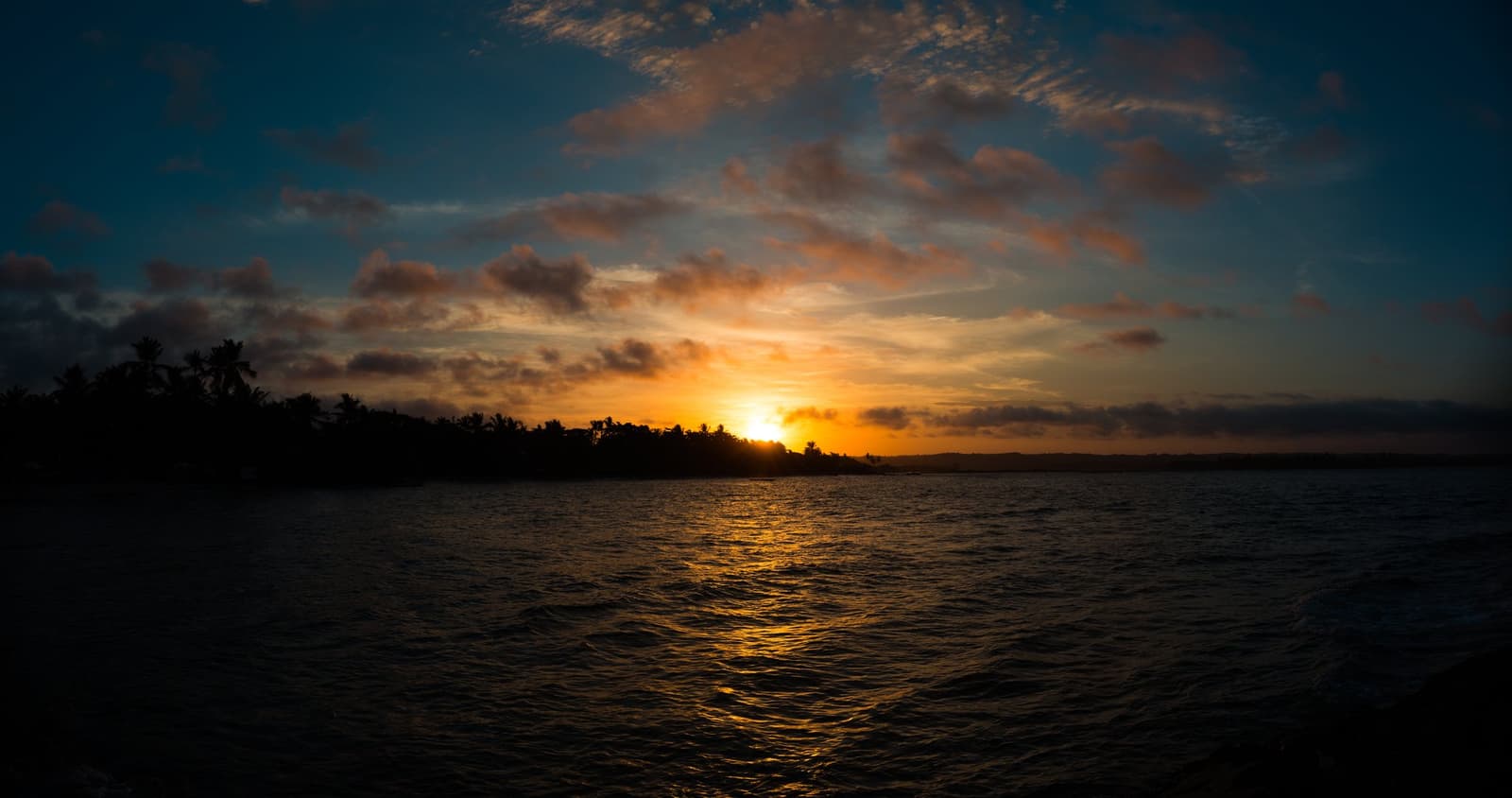 O mar recolhe, o céu abre. Pôr do sol dourado refletido sobre a água calma do oceano, com a silhueta escura de um litoral coberto de coqueiros no horizonte, visto de dentro d'água