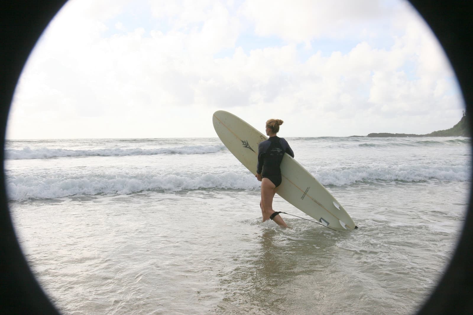 Prancha na mão, olho no mar, cabeça na próxima onda. Photo: Picasa Uma mulher de roupa de neoprene preta carrega uma prancha longa e entra no mar durante uma aula na escola de surf de Itacaré, fotografada com efeito de vinheta circular e ondas quebrando ao fundo