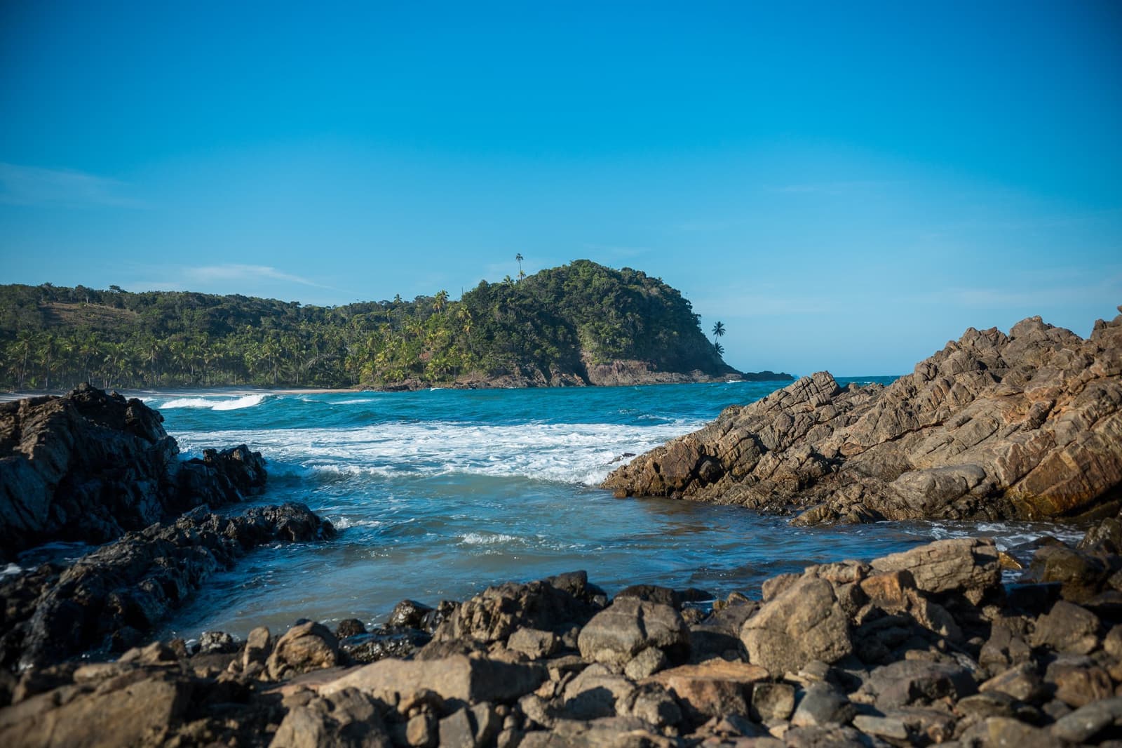 Pedras, mata fechada, água morna, a sala de aula fica aqui fora. Rochas escuras e recortadas emolduram uma enseada no litoral da escola de surf em Itacaré, com ondas turquesa avançando em direção a uma praia de areia sob um promontório coberto de mata e céu azul límpido