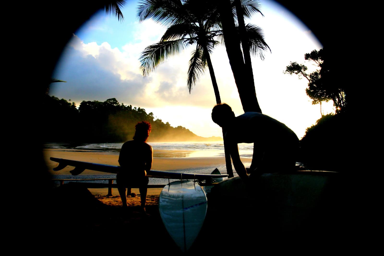 Último raio de luz, pranchas prontas, e o mar ainda tem onda pra dar. Photo: Picasa Dois surfistas em silhueta contra um pôr do sol dourado em uma praia tropical da escola de surf em Itacaré, passando cera e verificando longboards ao lado de coqueiros com morros cobertos de mata ao fundo